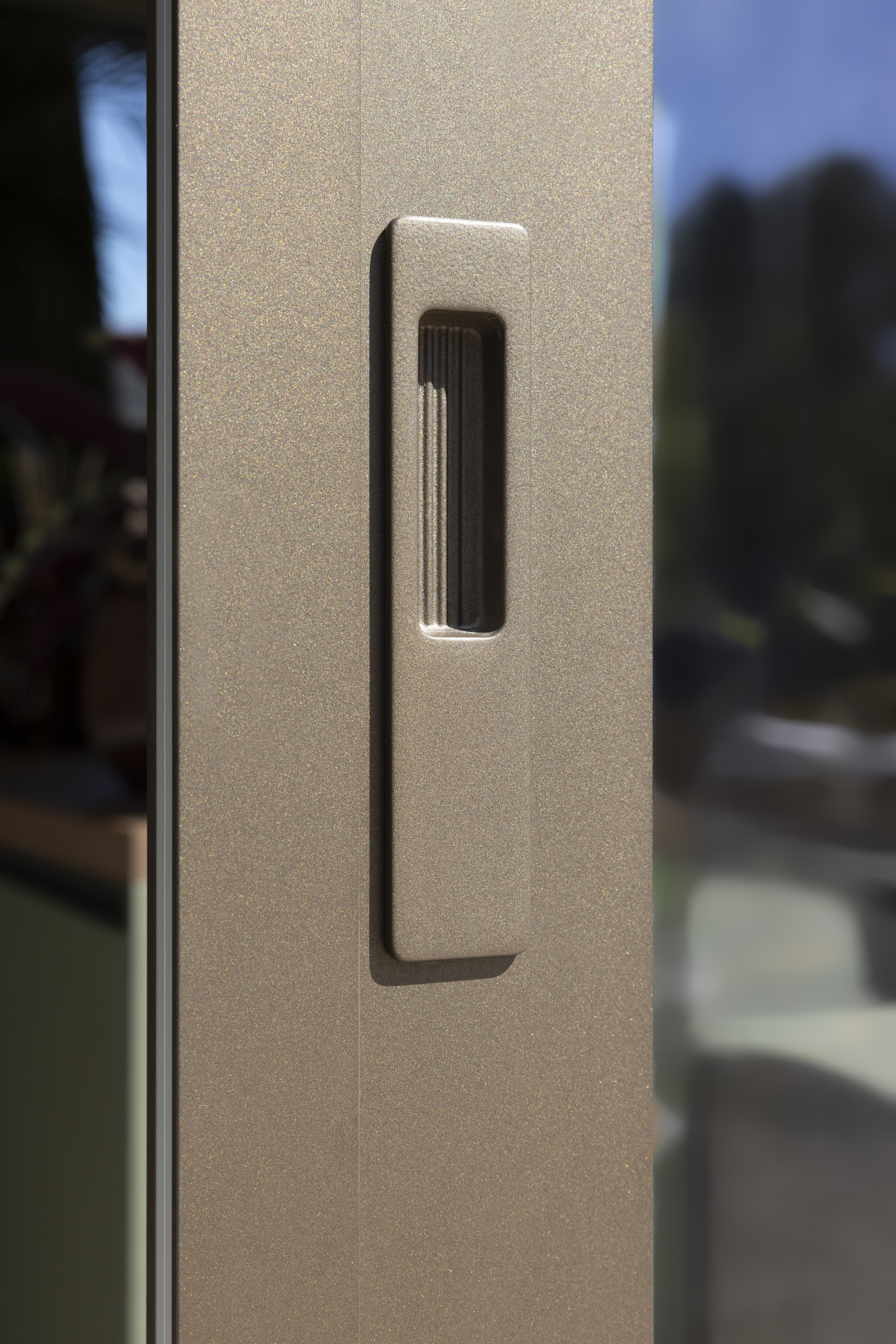 Close-up van een bronzen geanodiseerd aluminiumprofiel van een ALU Visoglide Monorail-schuifraam, met een verticale verzonken handgreep en glas op de achtergrond.