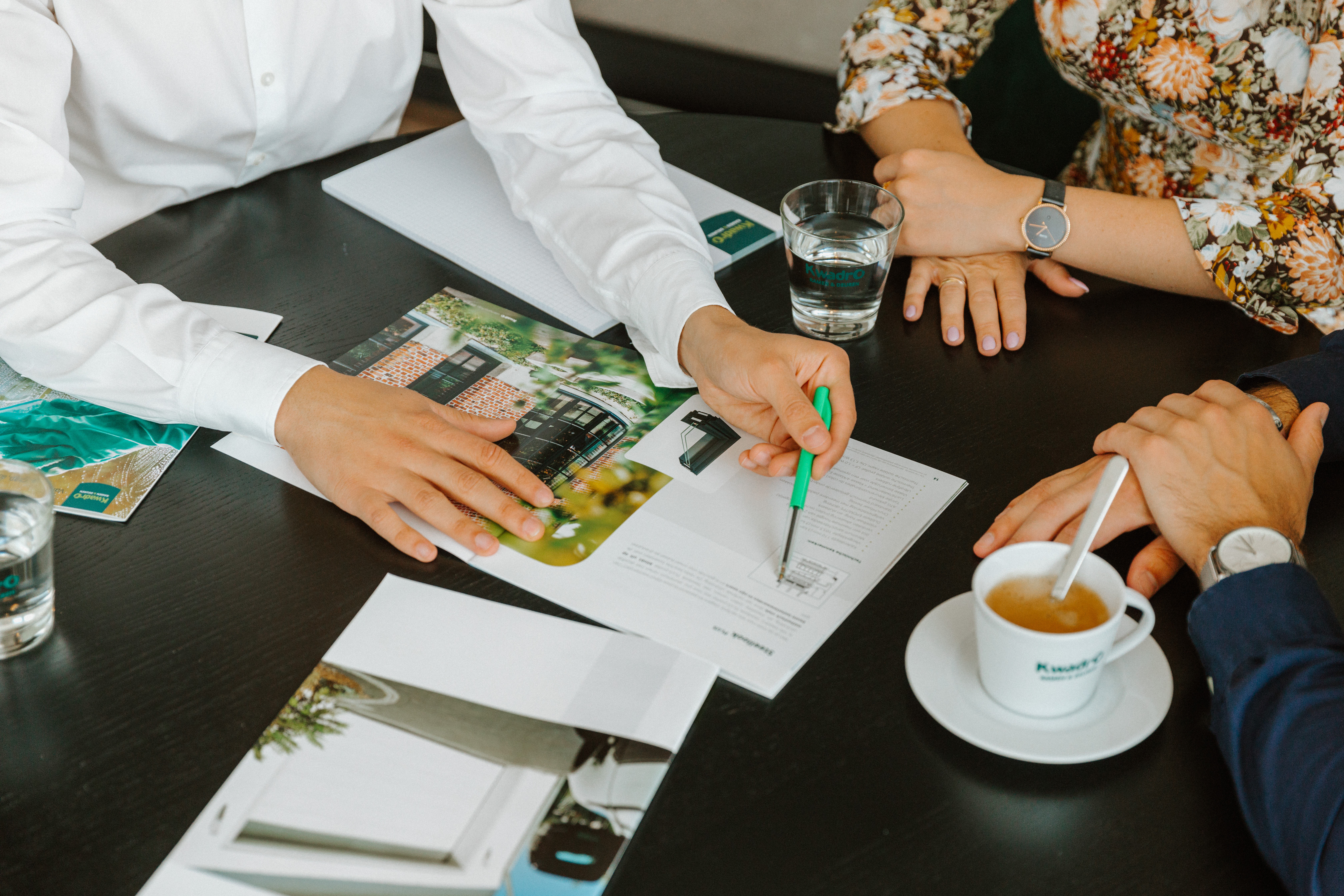 Plusieurs personnes examinent des brochures et des documents autour d’une table ; l’une d’elles écrit, avec du café et de l’eau à côté.