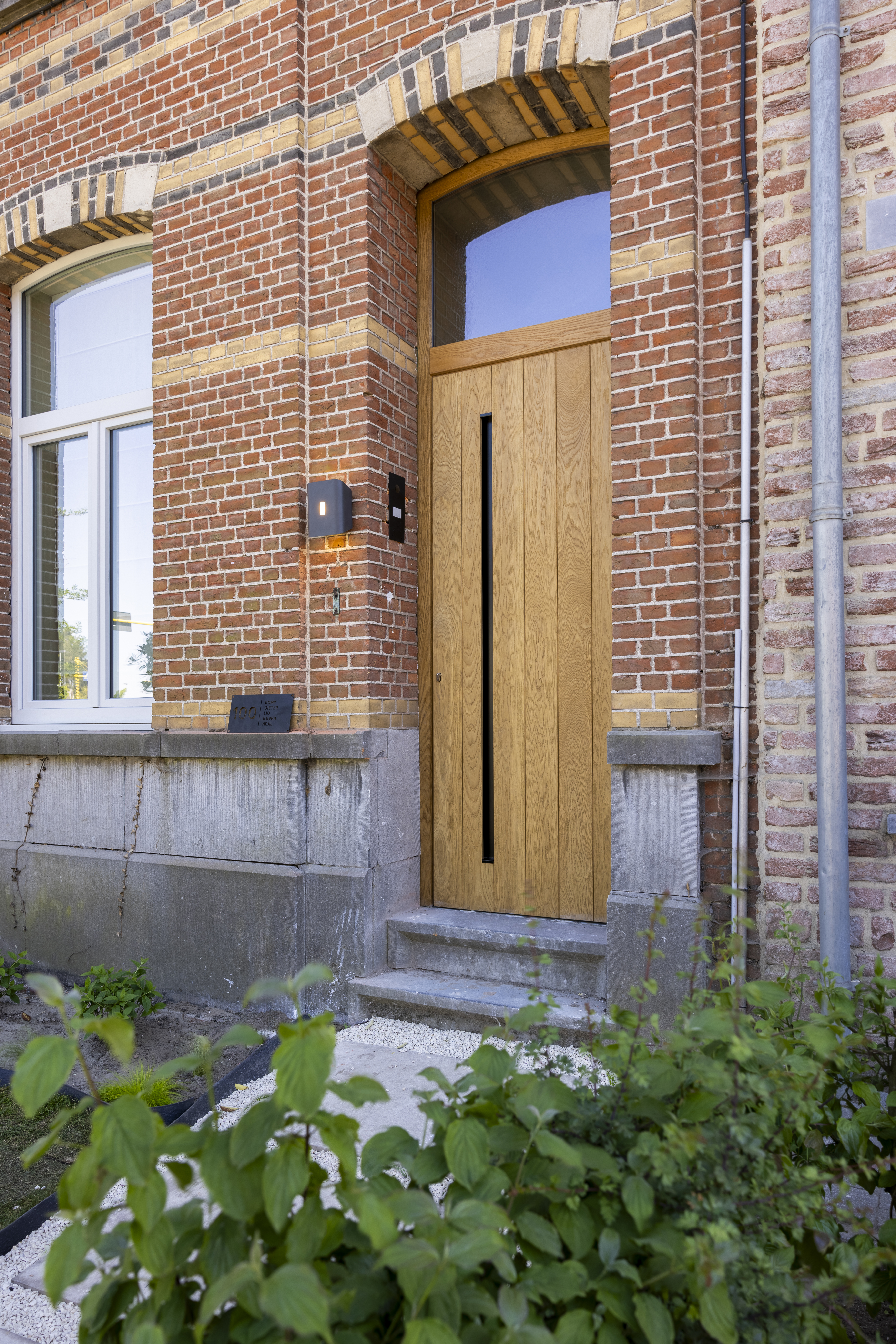 Eikenhouten deur met bovenlicht in een bakstenen gevel, naast een hoog raam en een kleine voortuin.