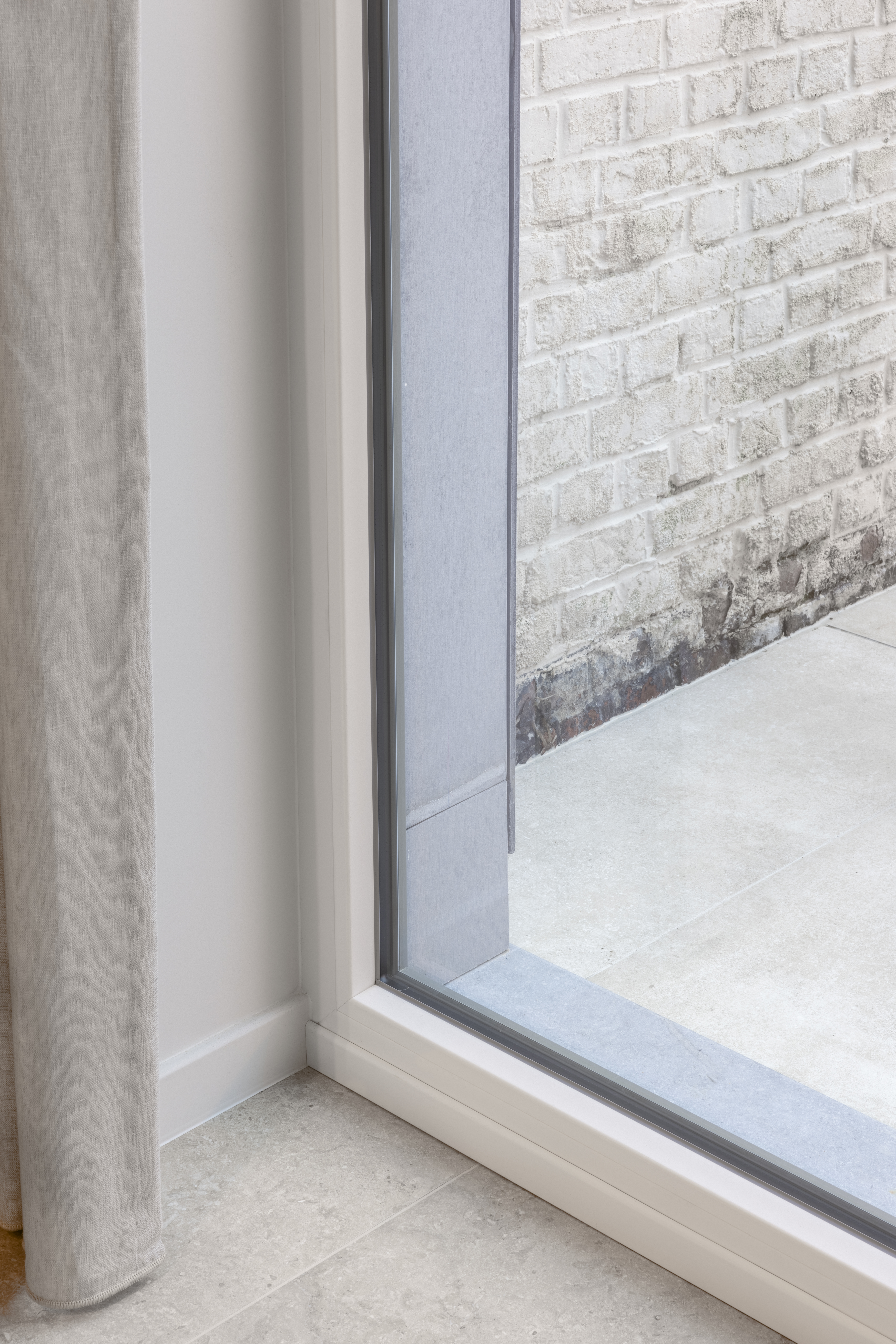 Modern vast raam met wit raamprofiel in een gerenoveerde gesloten bebouwing; buiten zijn een lichte bakstenen muur en een lichte vloer zichtbaar.