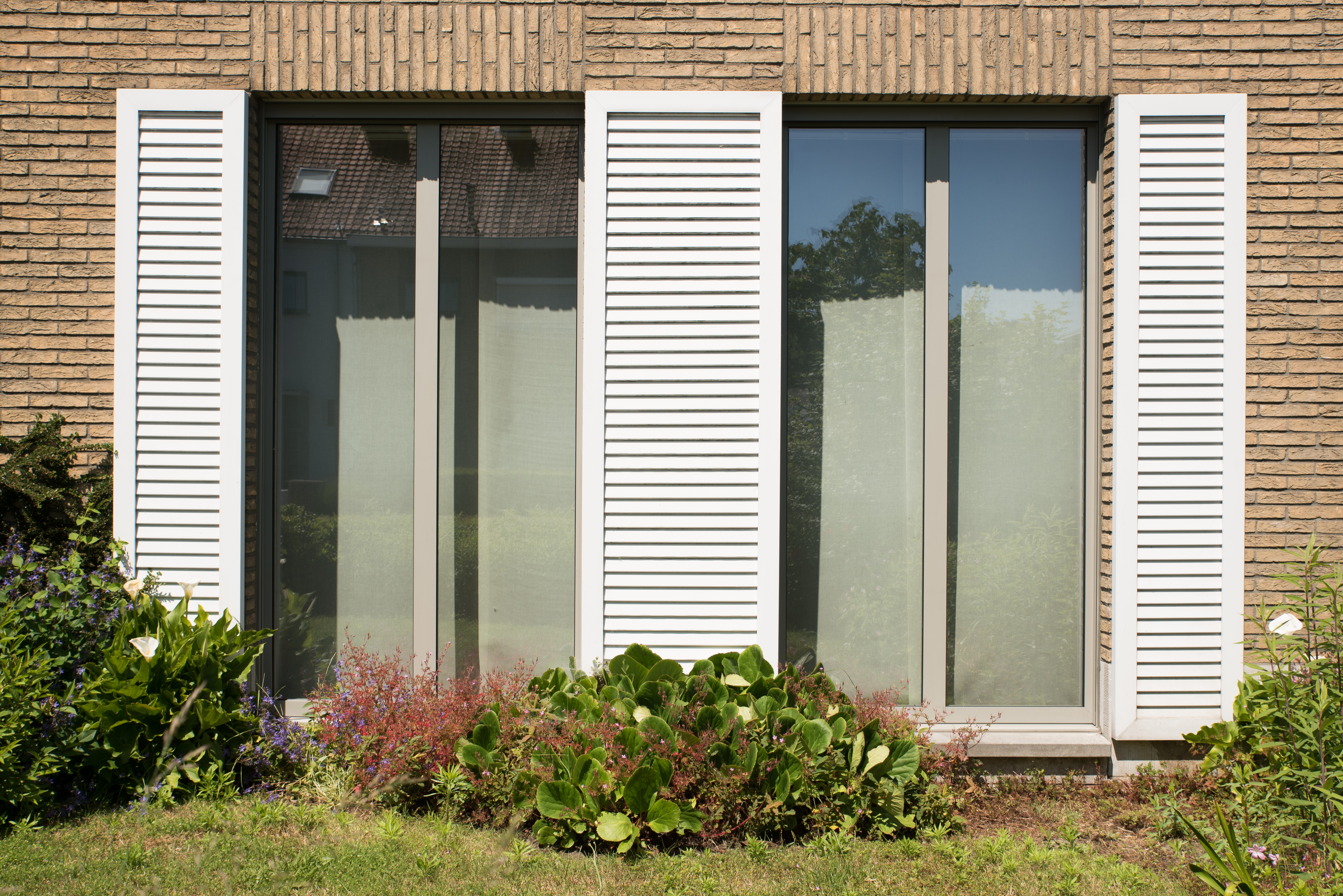 Gerestaureerde bakstenen gevel van een driegevelwoning met twee hoge ramen, witte luiken en een beplante voortuin.