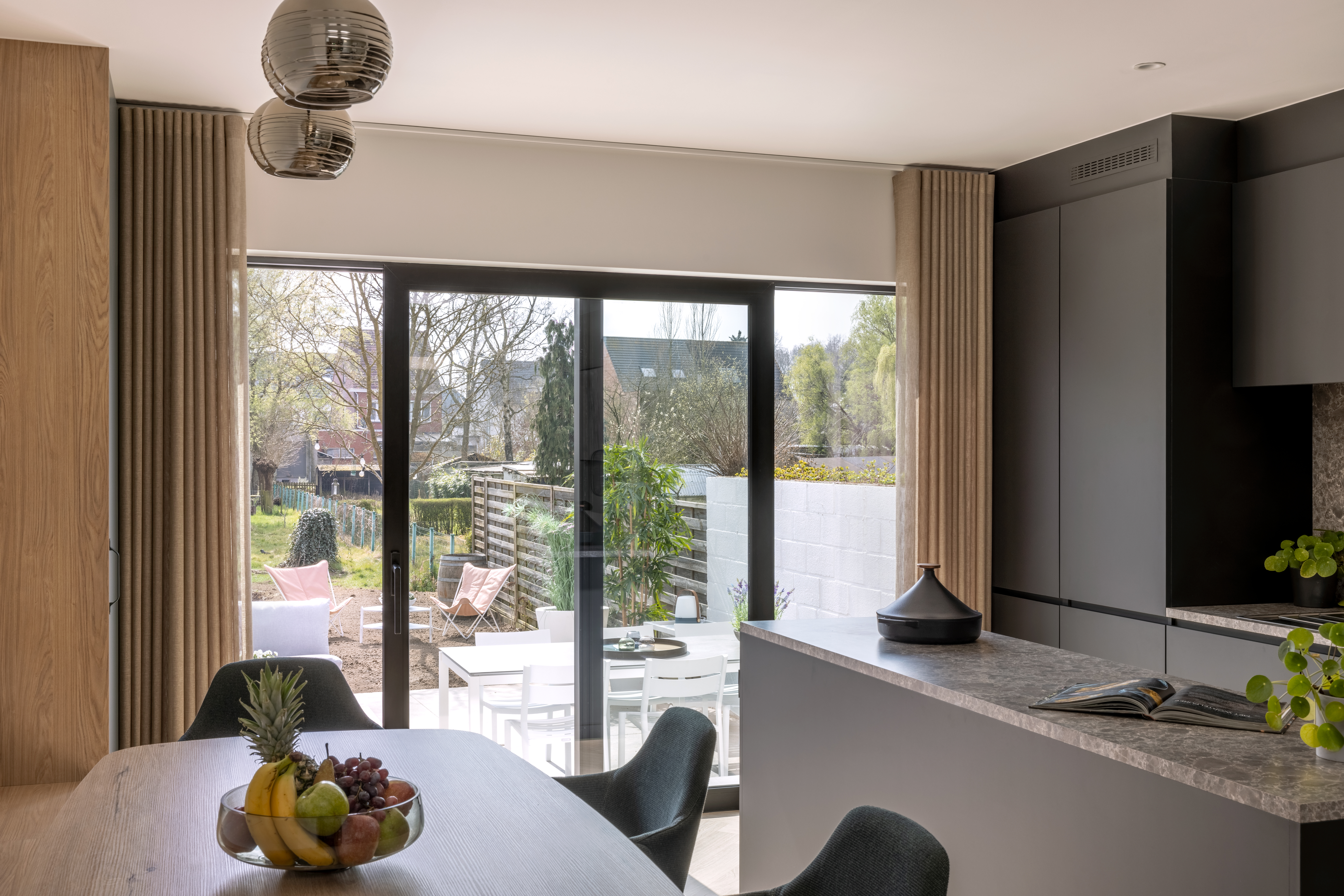 Lichte keuken met eethoek en grote zwarte glazen schuiframen naar een terras met zicht op de tuin, aangevuld met houten accenten en een keukeneiland.