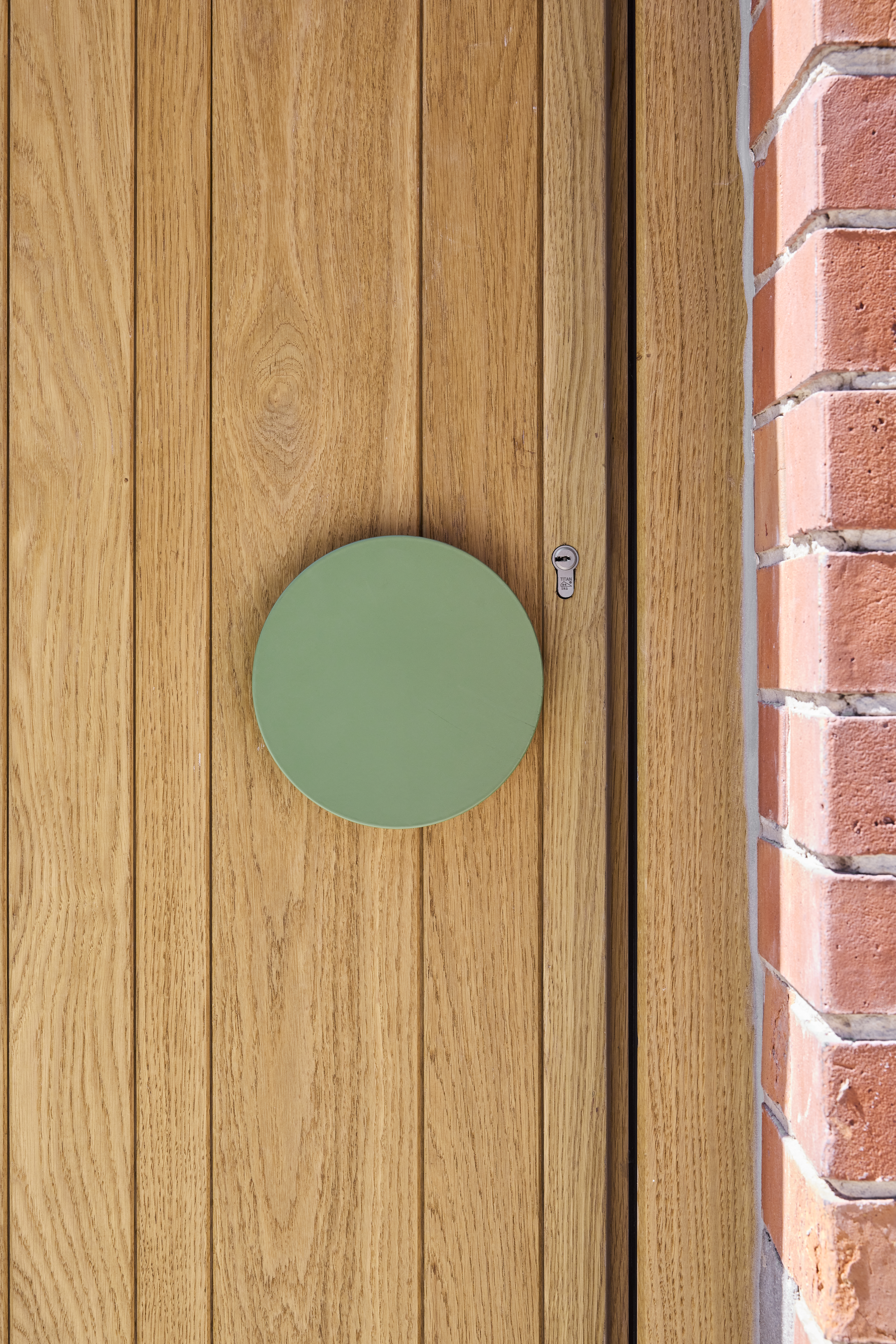 Close-up van een eikenhouten deur naast een rode bakstenen gevel, met een verticale houtstructuur en een rond groen detail.