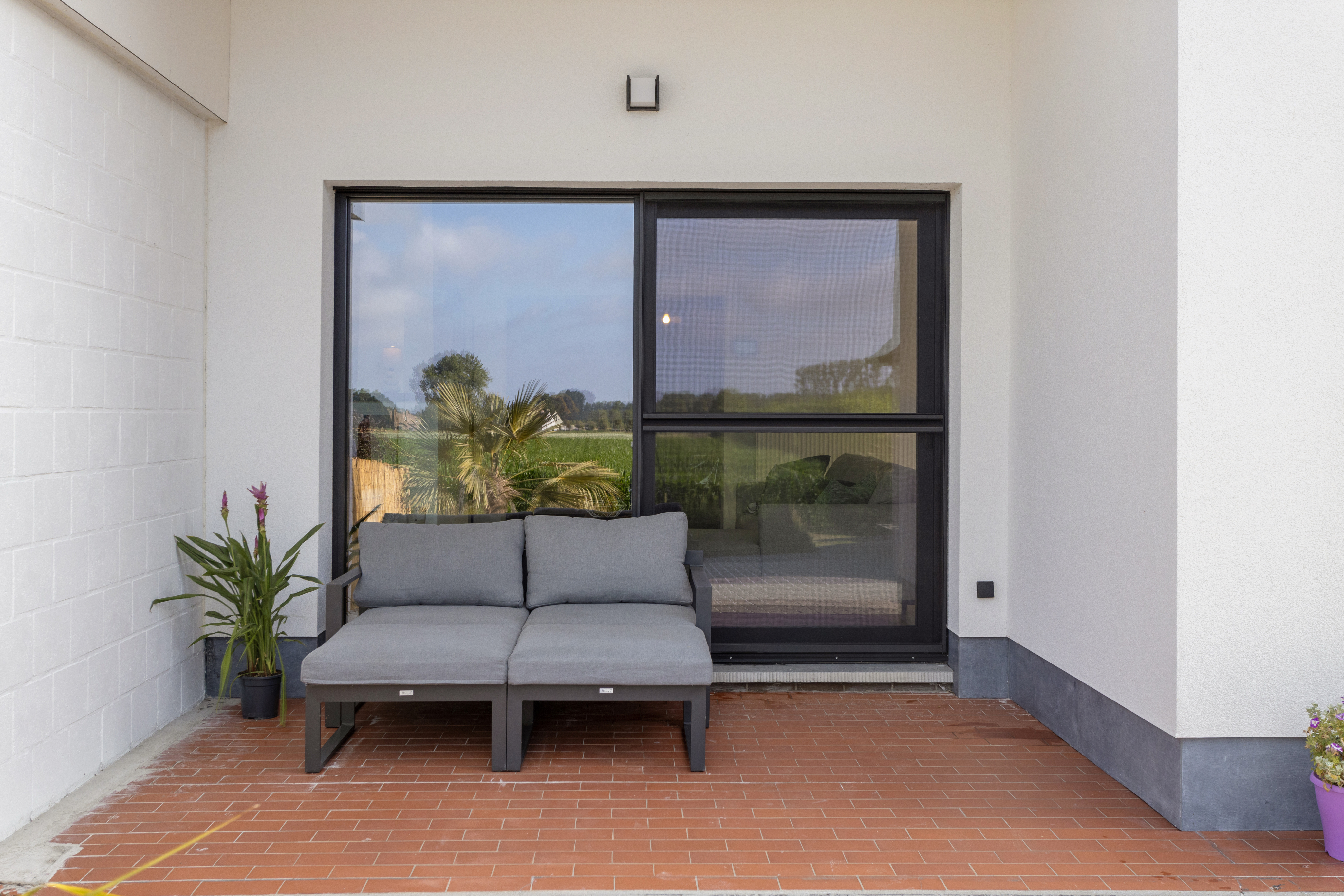 Modern schuifvliegenraam in matzwart aluminium aan een overdekt terras, met zicht op de tuin en een loungebank tegen de gevel.
