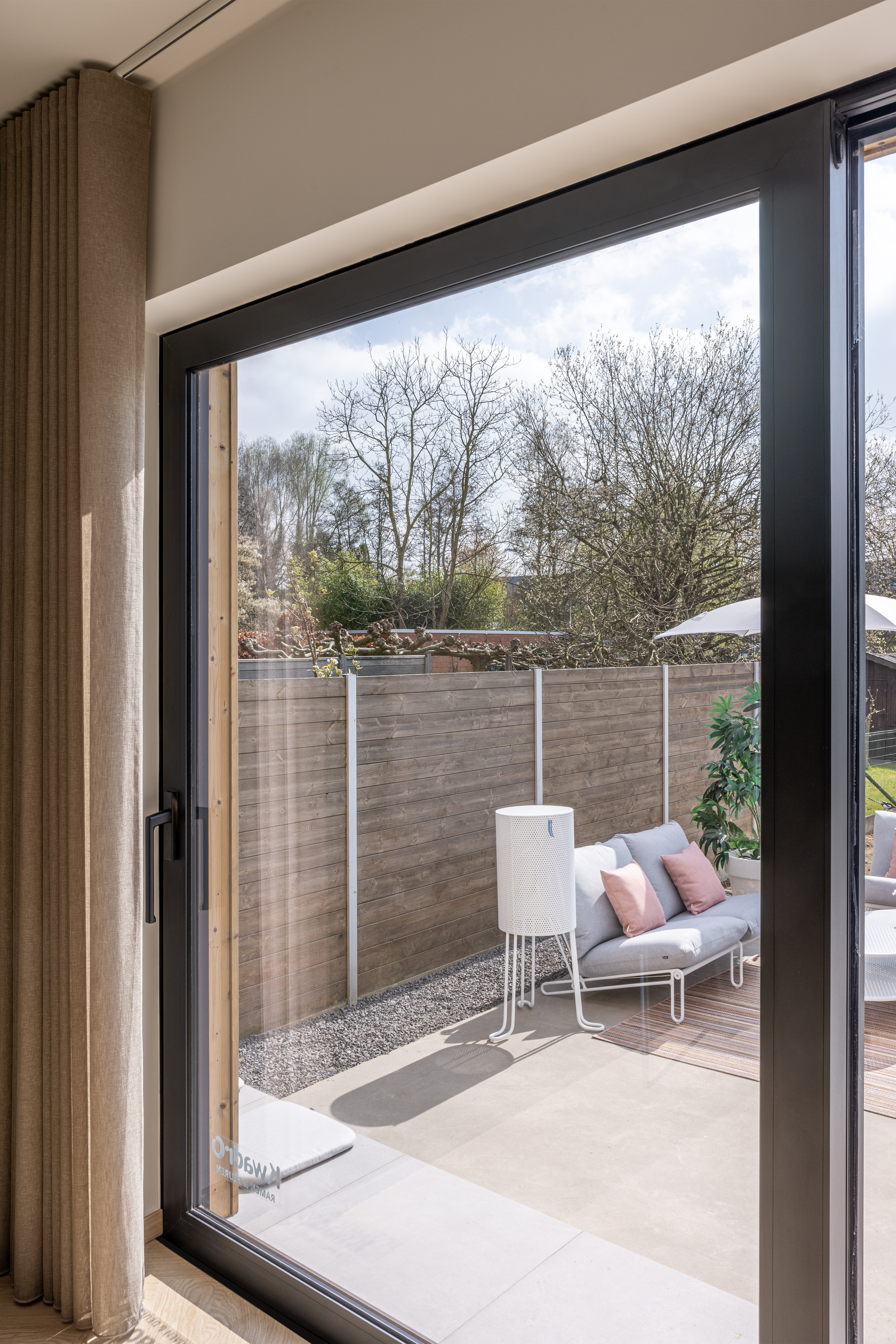 Grote glazen raamopening met grijze profielen biedt uitzicht op een betegeld terras met tuinmeubelen en een omheinde tuin.