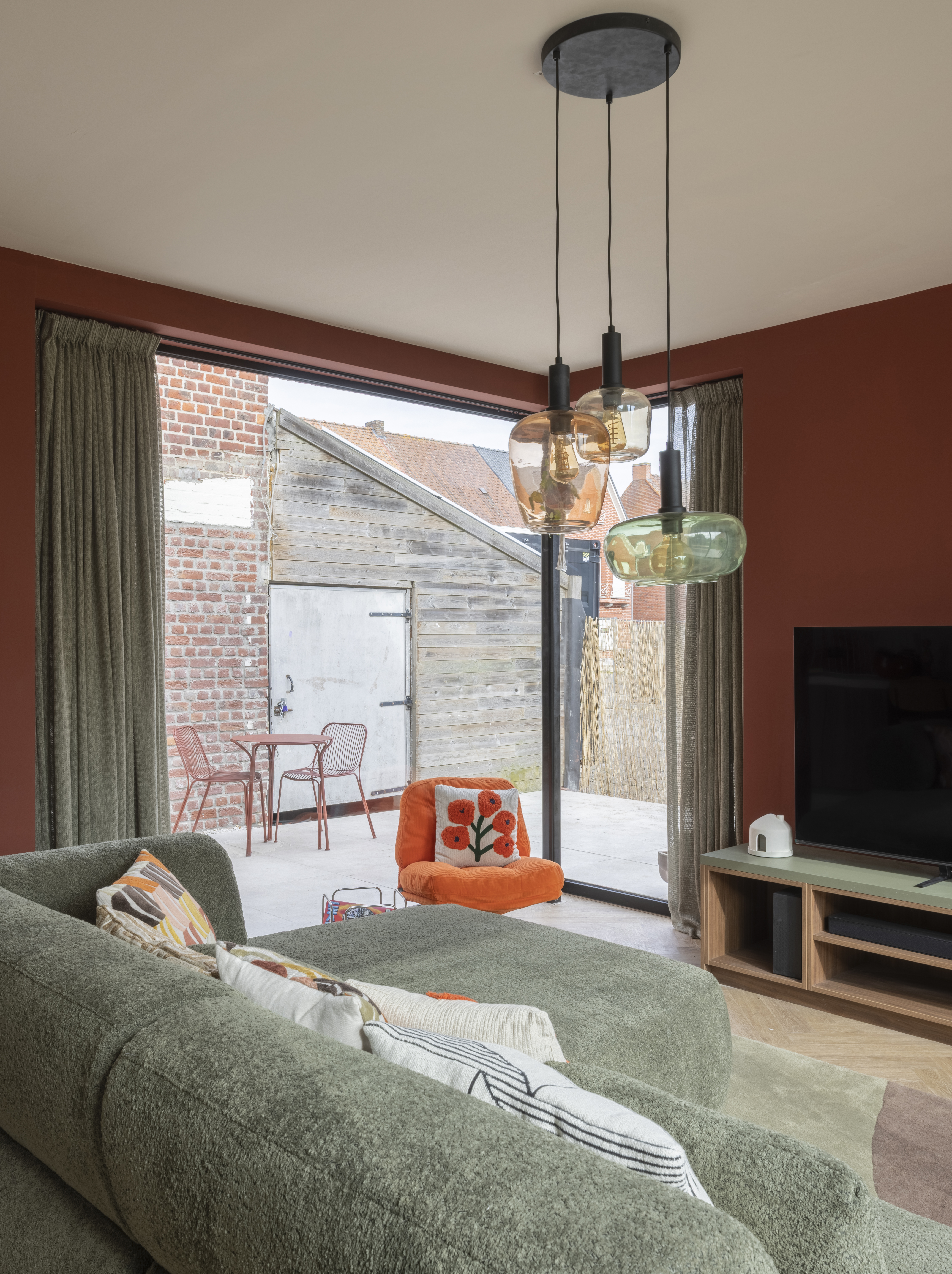 Woonkamer met een groot vast raam in diepzwart, grijze gordijnen, een zetel, een televisie en zicht op een bakstenen gevel.