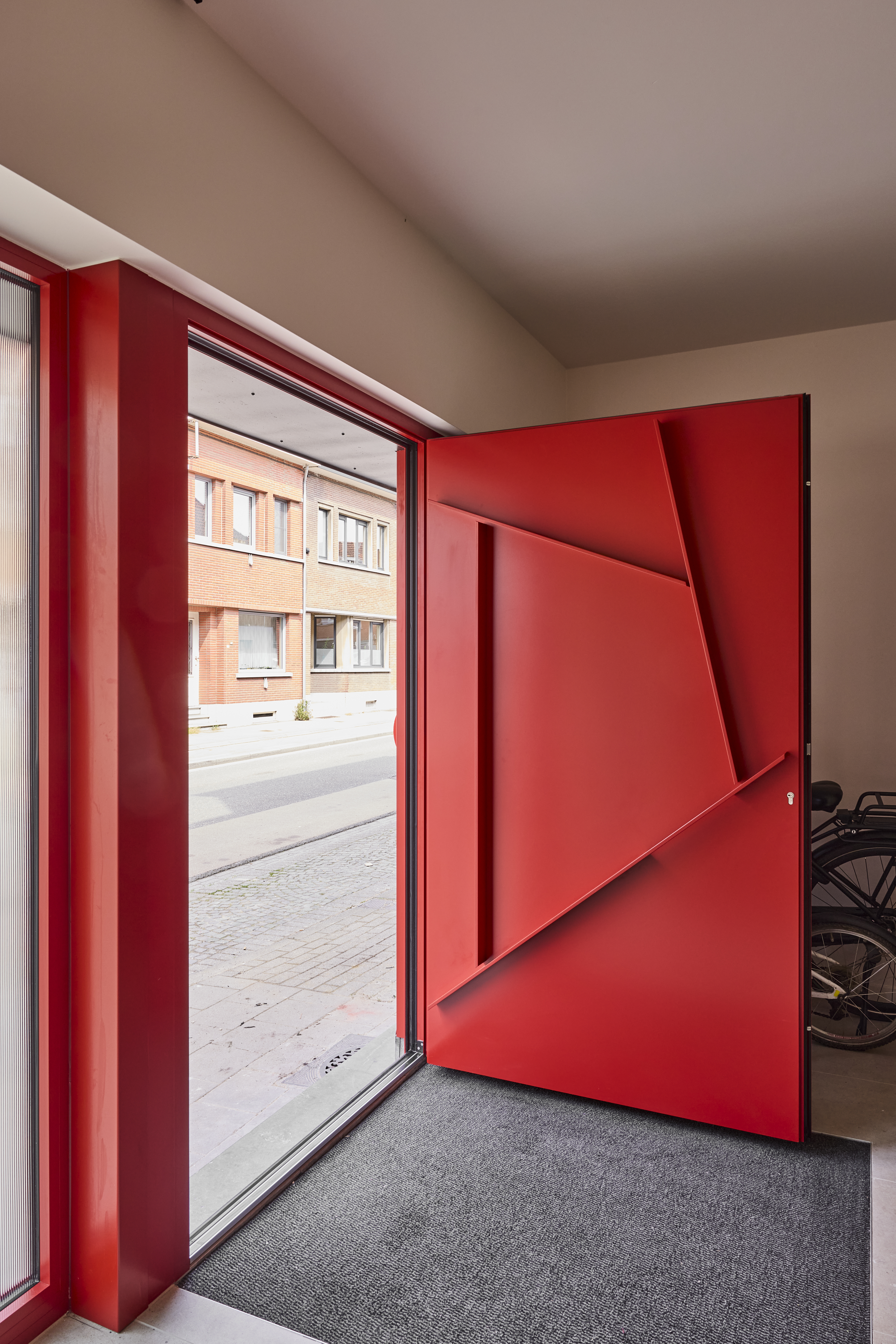 Rode aluminium­voordeur Komfort EVO Small met opvallend paneeldesign, geopend naast een smalle beglazing in een lichte inkomhal.