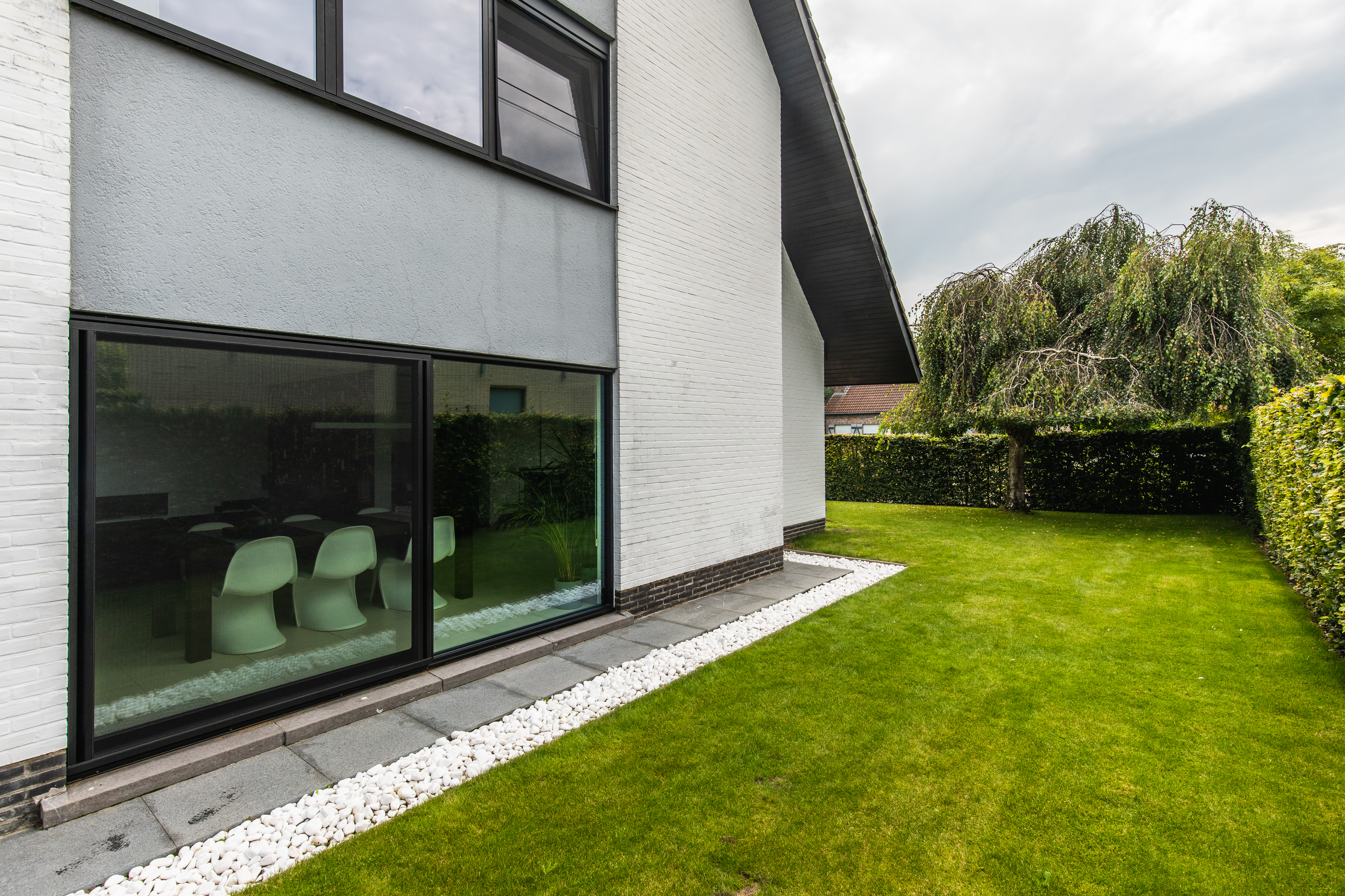 Moderne vrijstaande woning met zwart ALU Visoglide Monorail-schuifraam en schuifvliegenraam, aan een verzorgd gazon met haag.