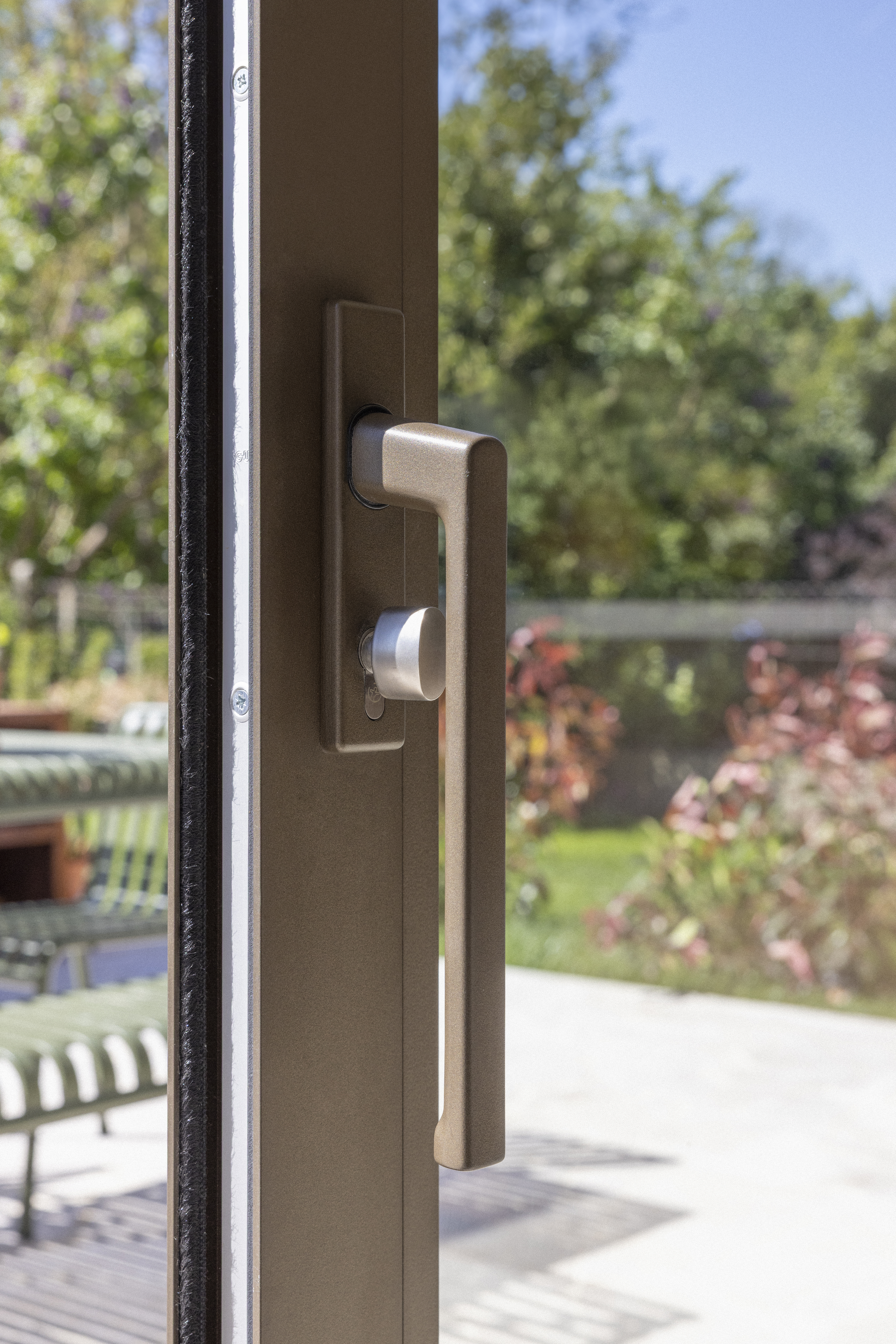 Close-up van een schuifraam in brons geanodiseerd aluminium, ALU Visoglide Monorail, met handgreep en uitzicht op een zonnige tuin.