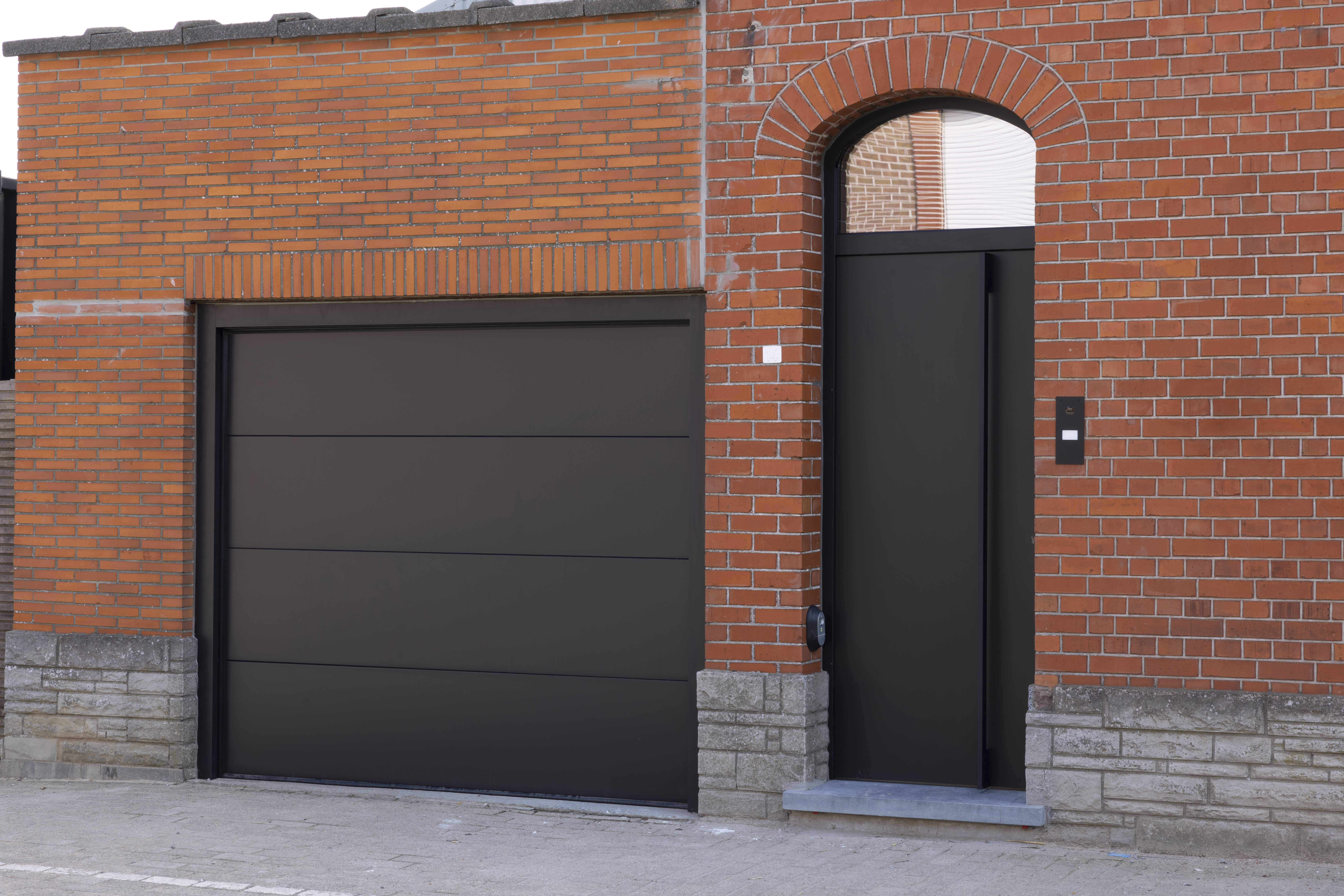 Porte d’entrée en PVC Komfort PLUS noir mat, avec imposte et panneau, à côté d’une porte de garage assortie dans une façade en briques rouges.
