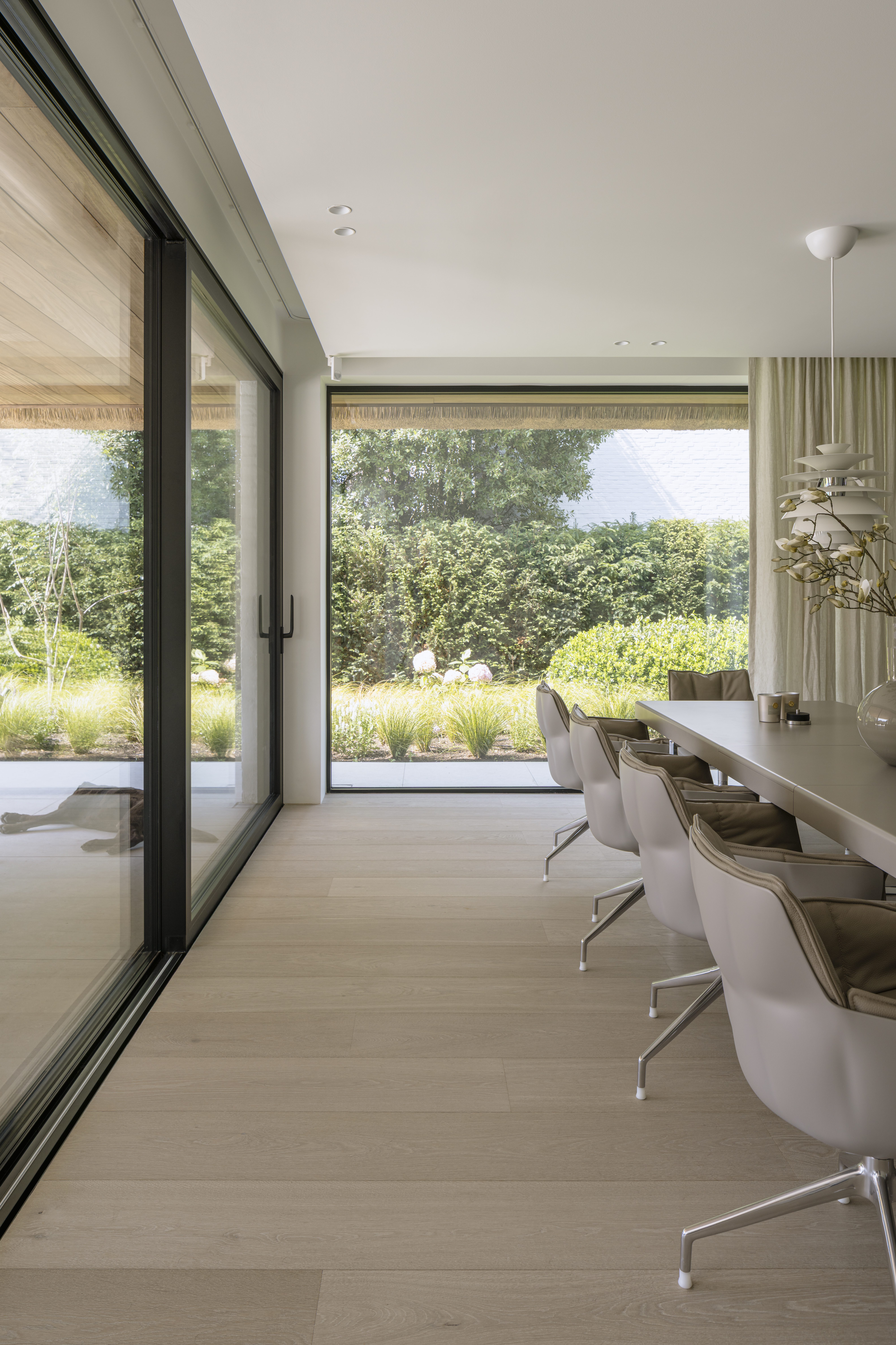 Licht interieur met een groot schuifraam, een lange eettafel en stoelen, met zicht op een groene tuin.