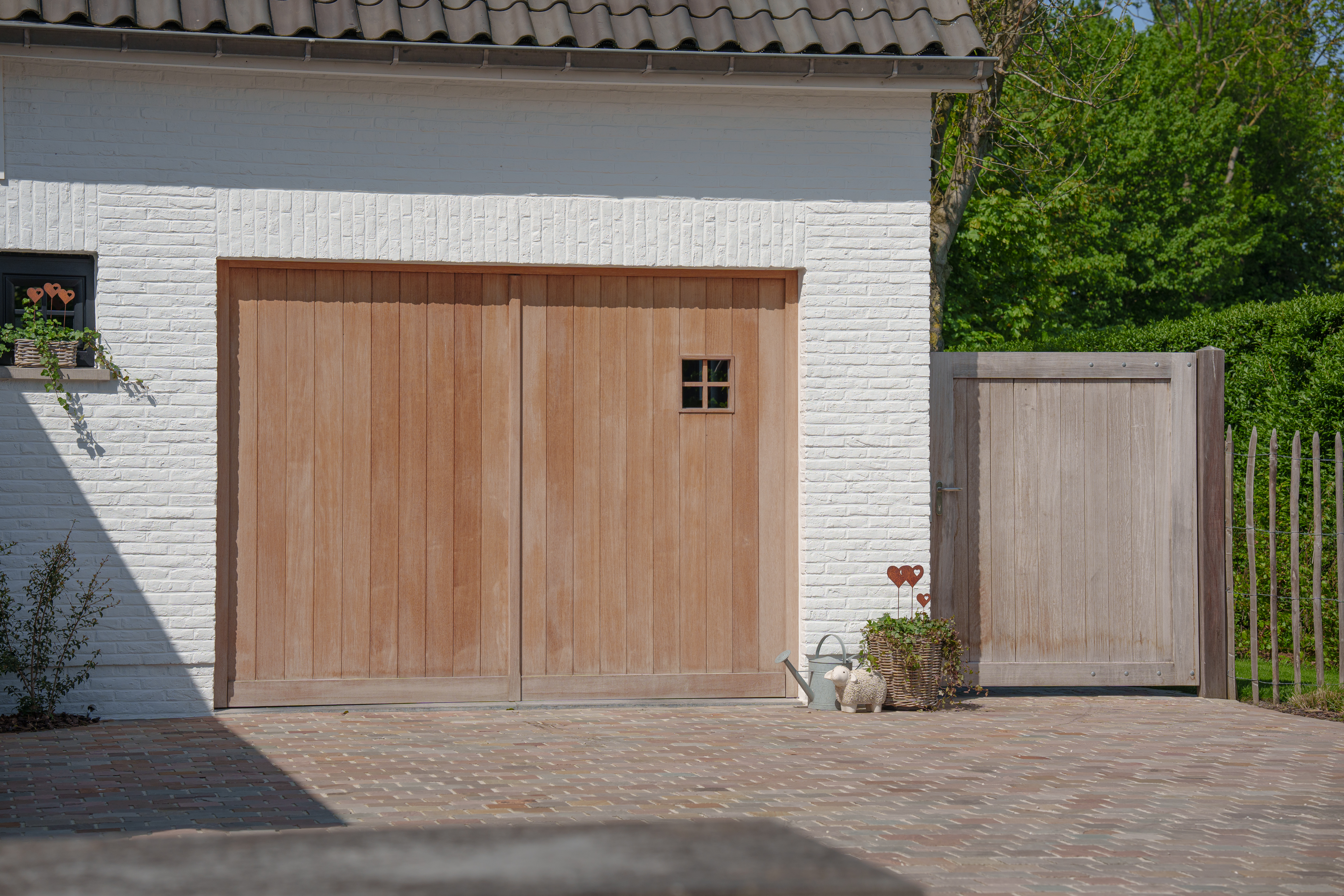 Houten dubbele garagepoort met een klein raam in een witgeschilderde bakstenen gevel, naast een bijpassende houten poort aan een gekasseide oprit.