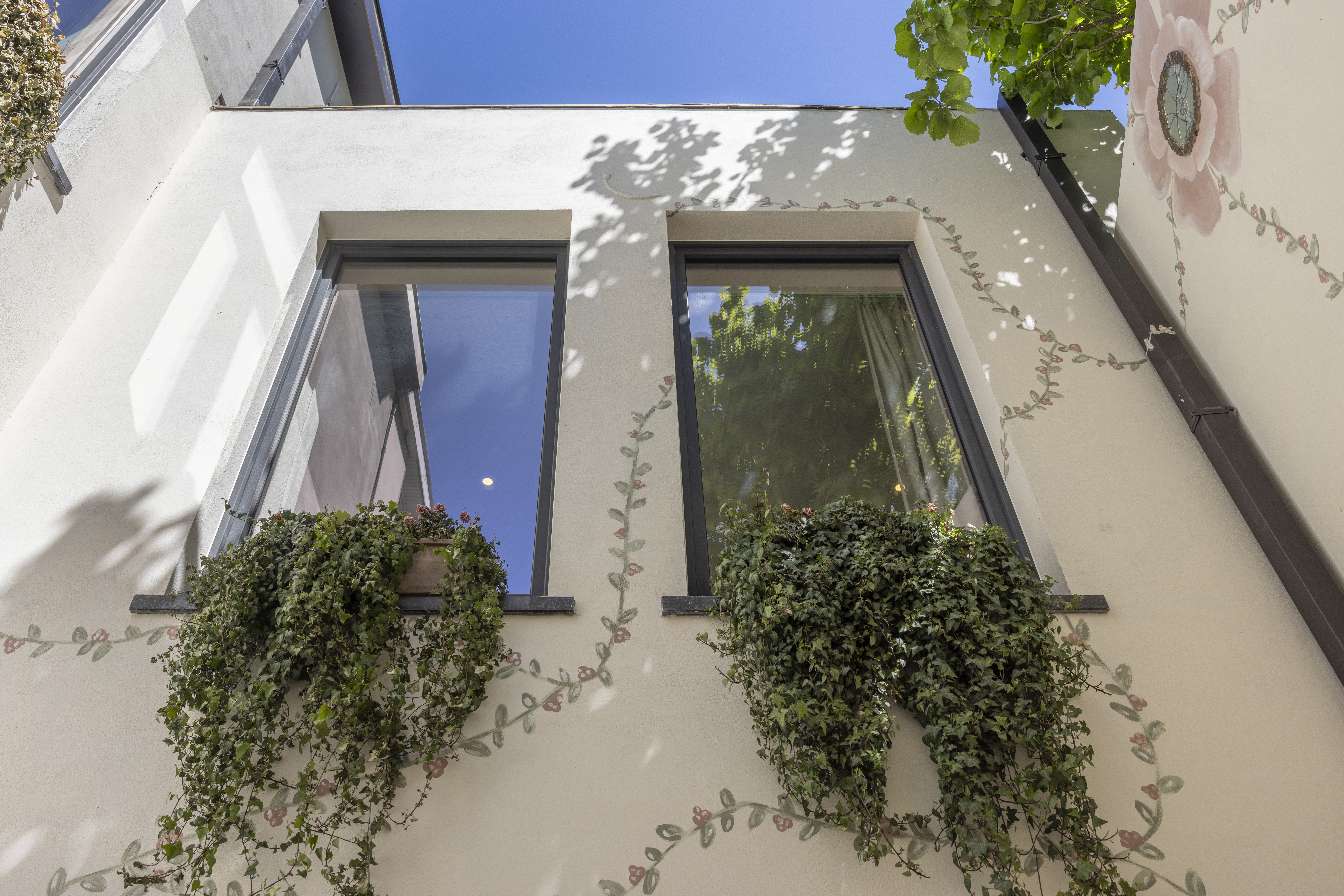 Twee mat bronzen draai-kipramen in de witte gevel van een lichte woning, deels omringd door hangende klimplanten.