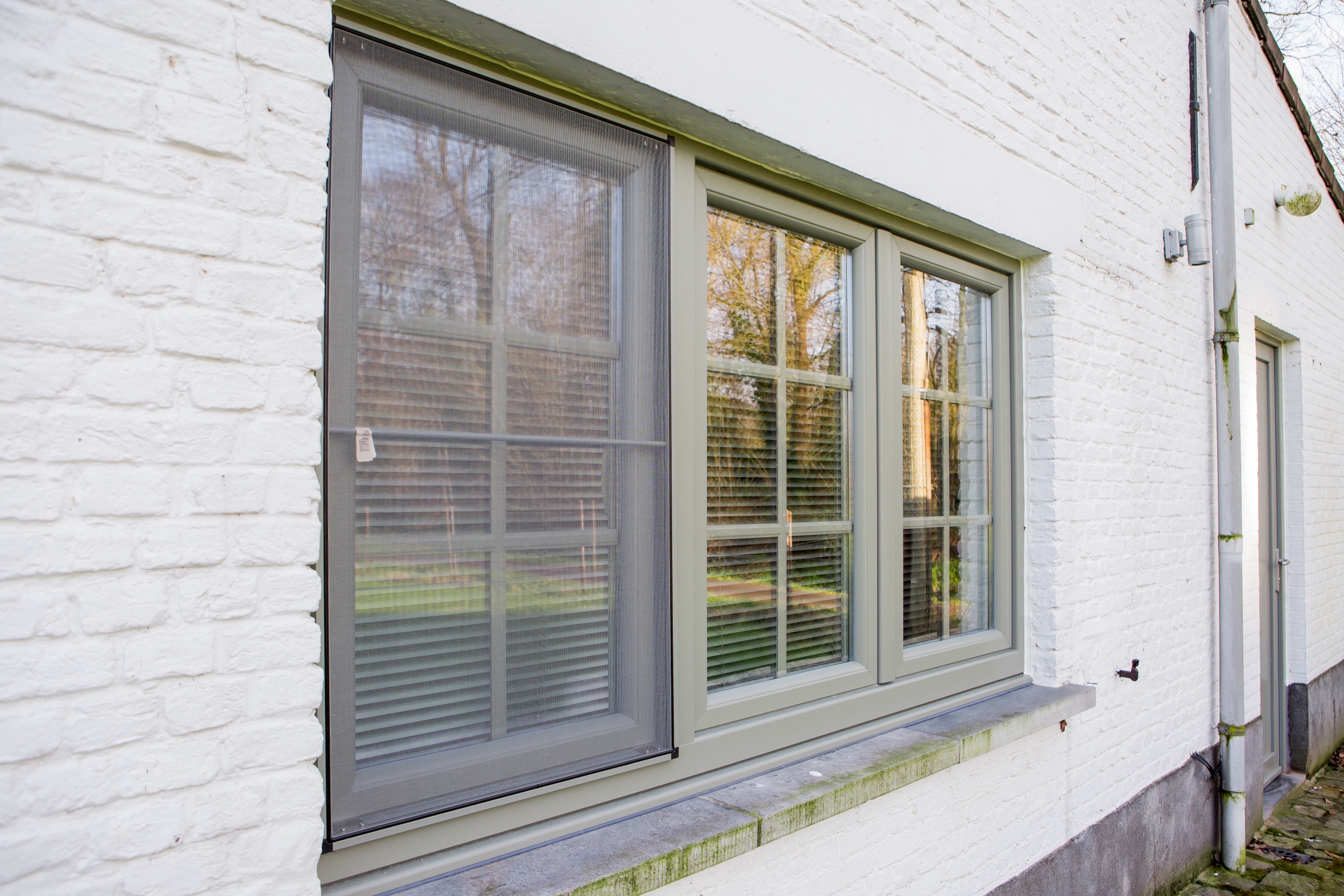 Betongrijs raam met drie verticale delen in een witte bakstenen gevel van een vrijstaande woning, passend bij een tijdloze renovatie.
