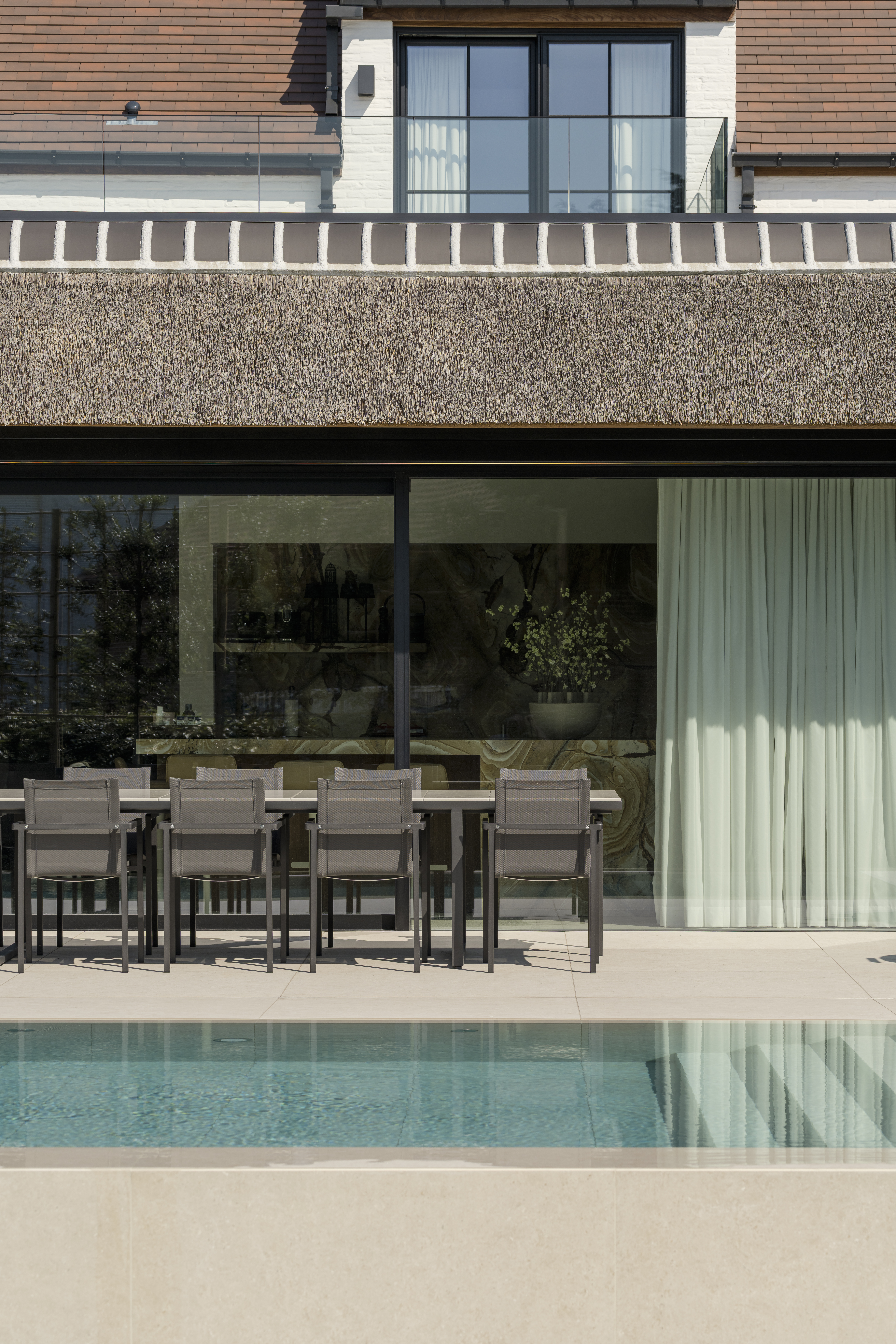 Moderne woning met een grote donkere raampartij aan het terras, zicht op het zwembad en een balkon met glazen balustrade.
