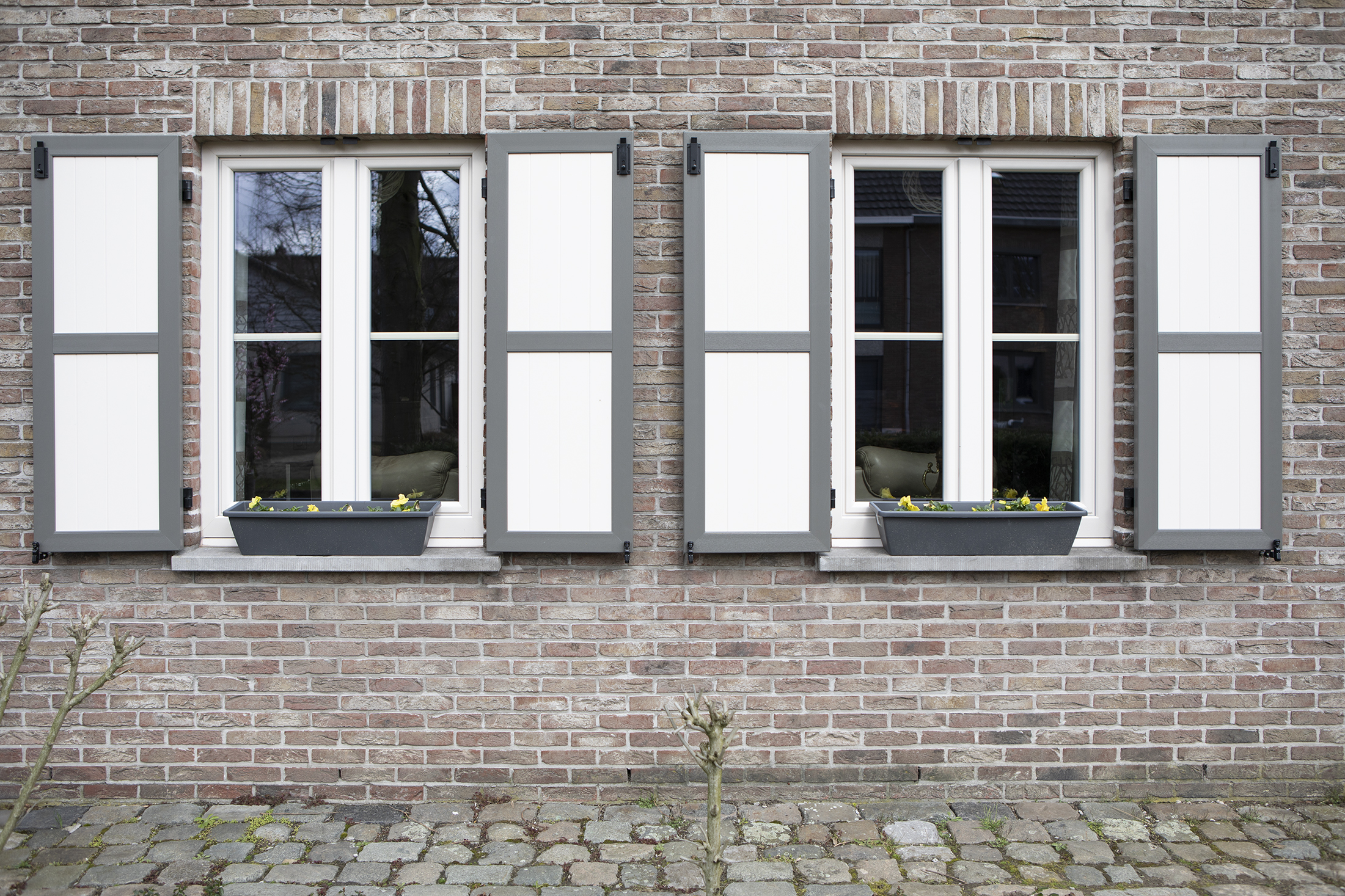 Bakstenen gevel met twee witte ramen en grijs-witte klapluiken, met bloembakken en een verharde strook onderaan.