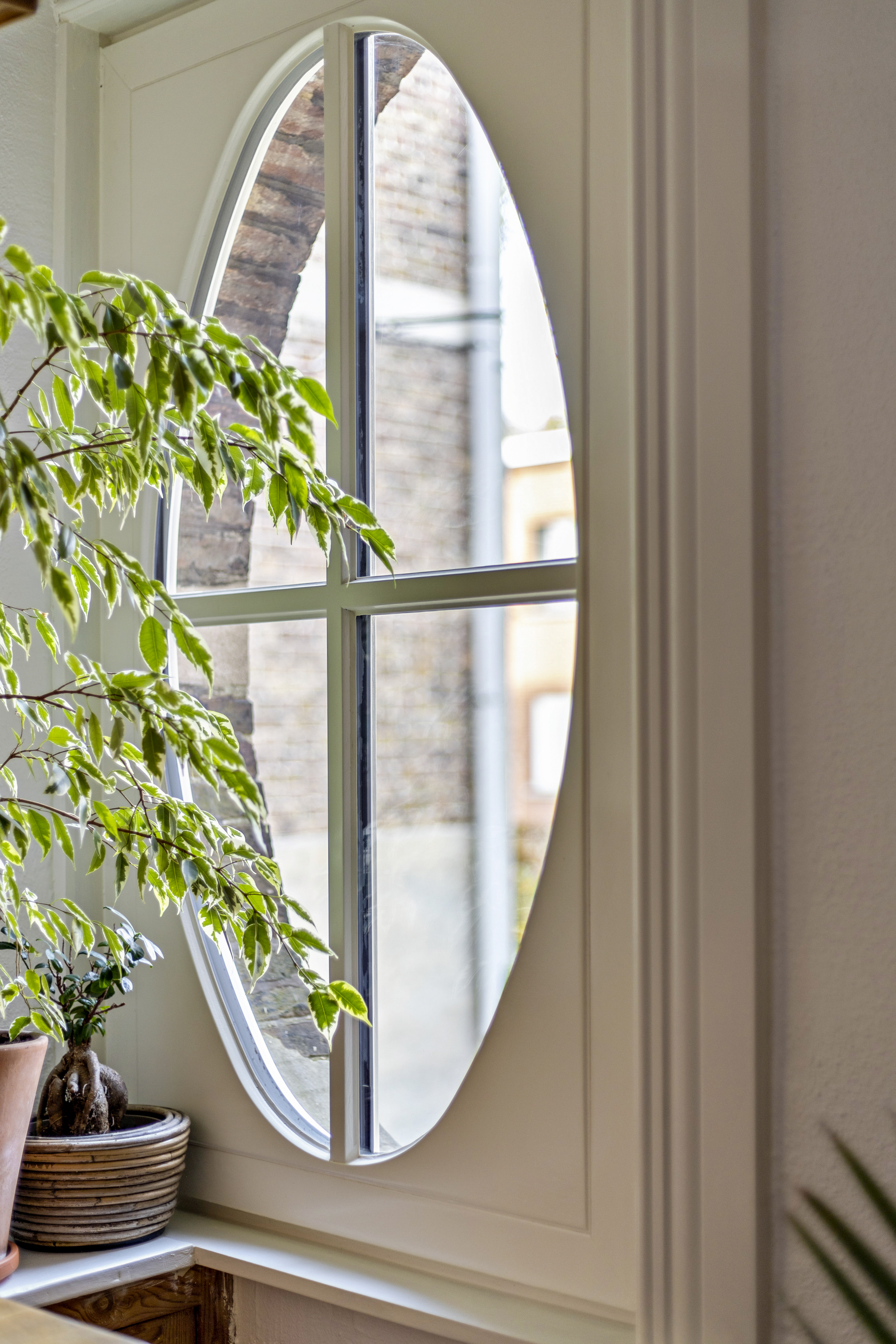 Afgelakt draai-kipraam met ovale vorm in een lichte wand, gedeeltelijk verscholen achter een kamerplant en met zicht op een bakstenen gevel.