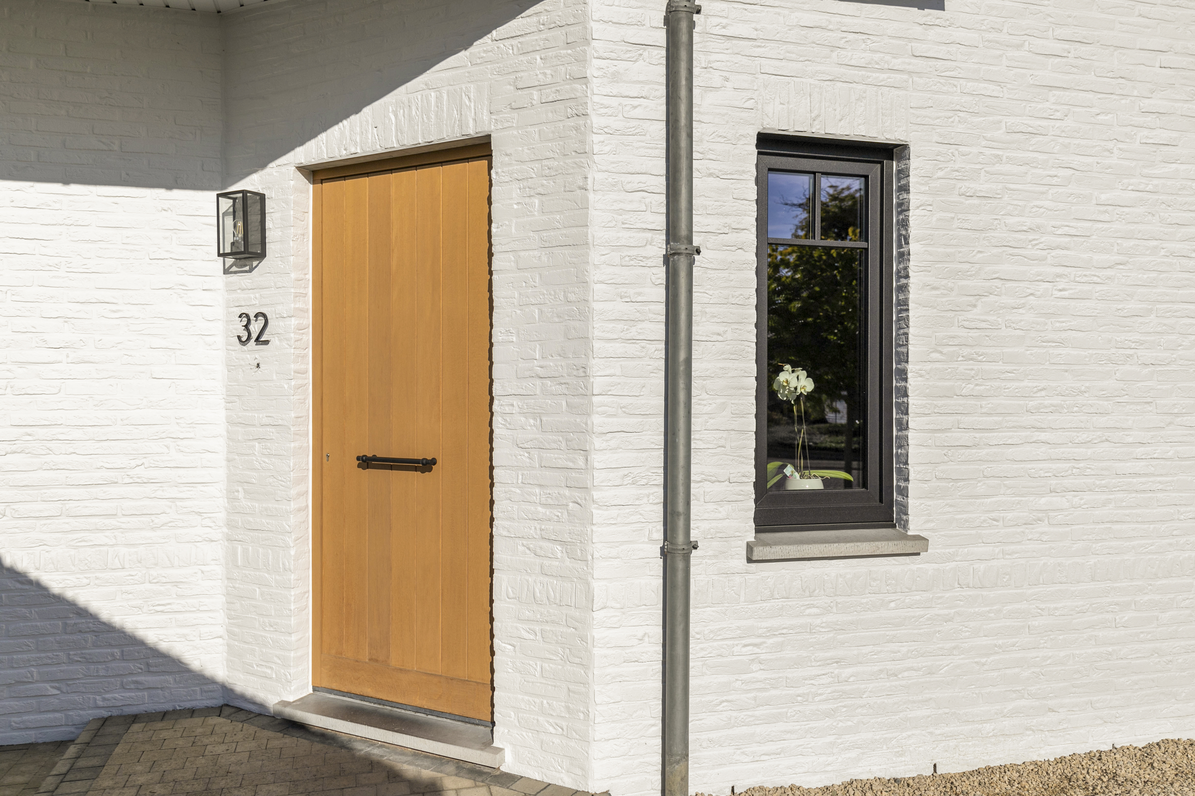 Witte bakstenen gevel met een zwart mat draai-kipraam in eik naast een houtkleurige voordeur, huisnummer 32 en regenpijp.
