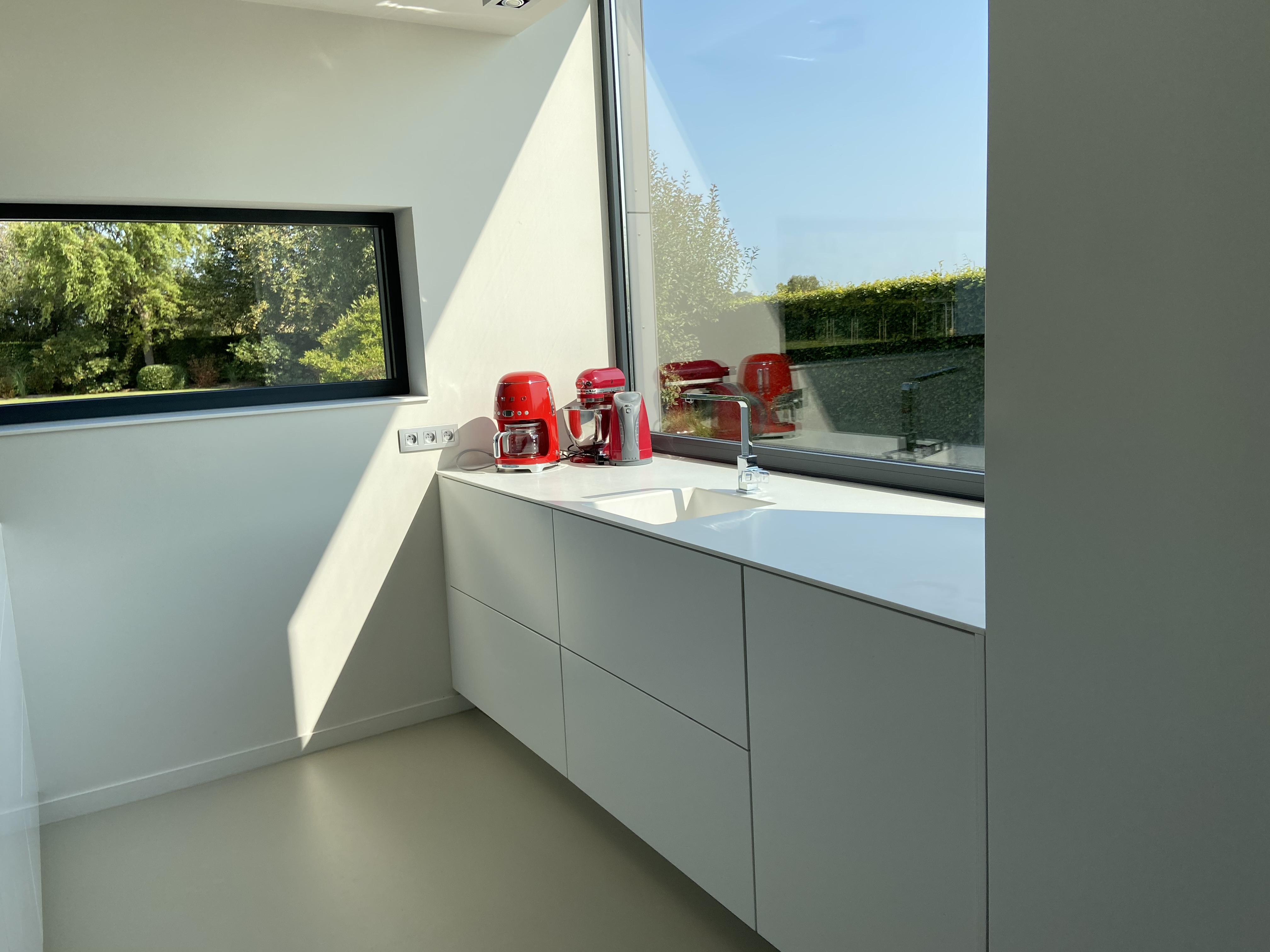 Lichte keuken in een open bebouwing met greeploze, lichtgrijze onderkasten, een hoekraam, een smal horizontaal raam en rode keukenaccessoires op het werkblad.