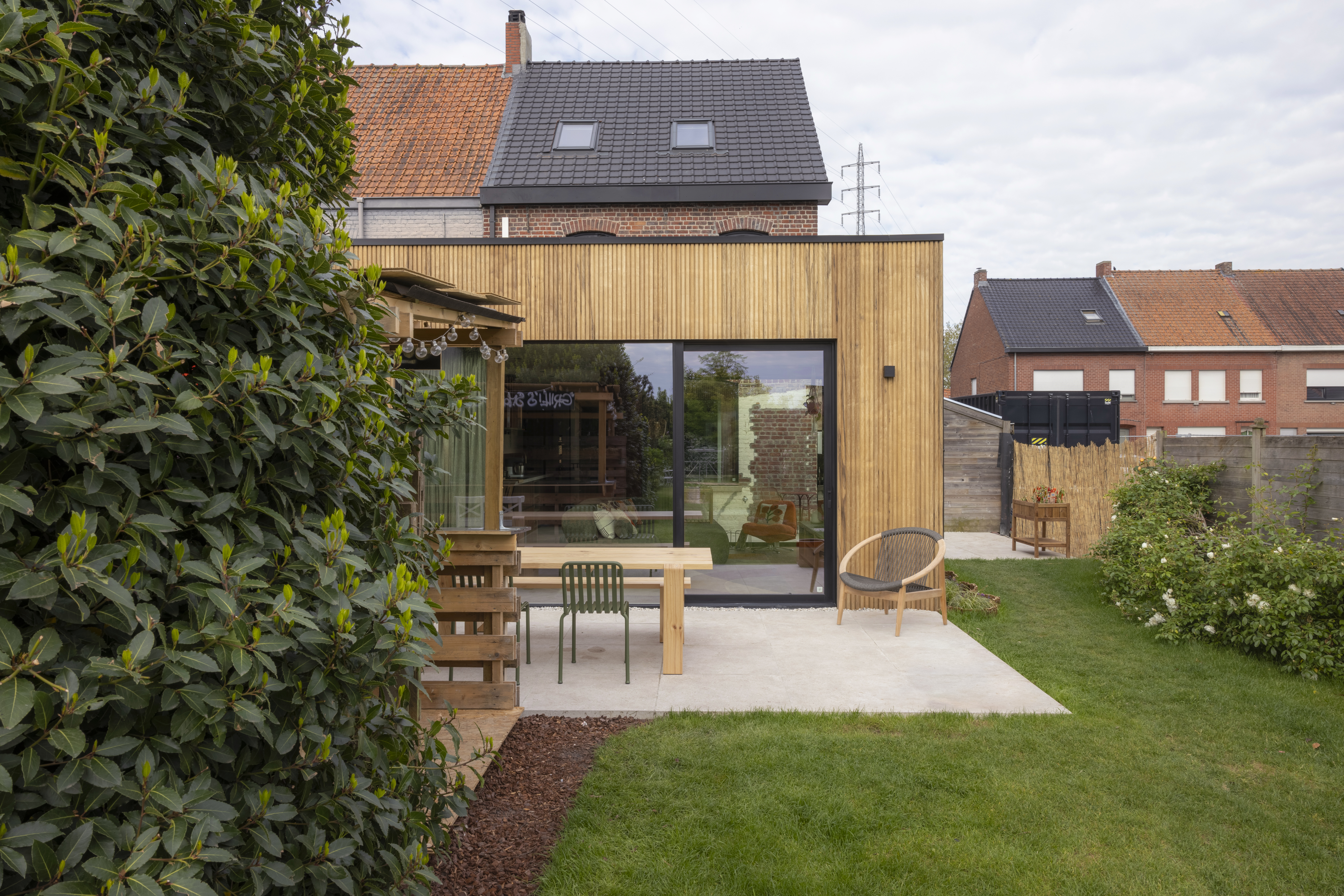 Houten aanbouw met een groot diepzwart ALU Visoglide Monorail-schuifraam, een terras met tuinmeubilair en aansluitend gazon.