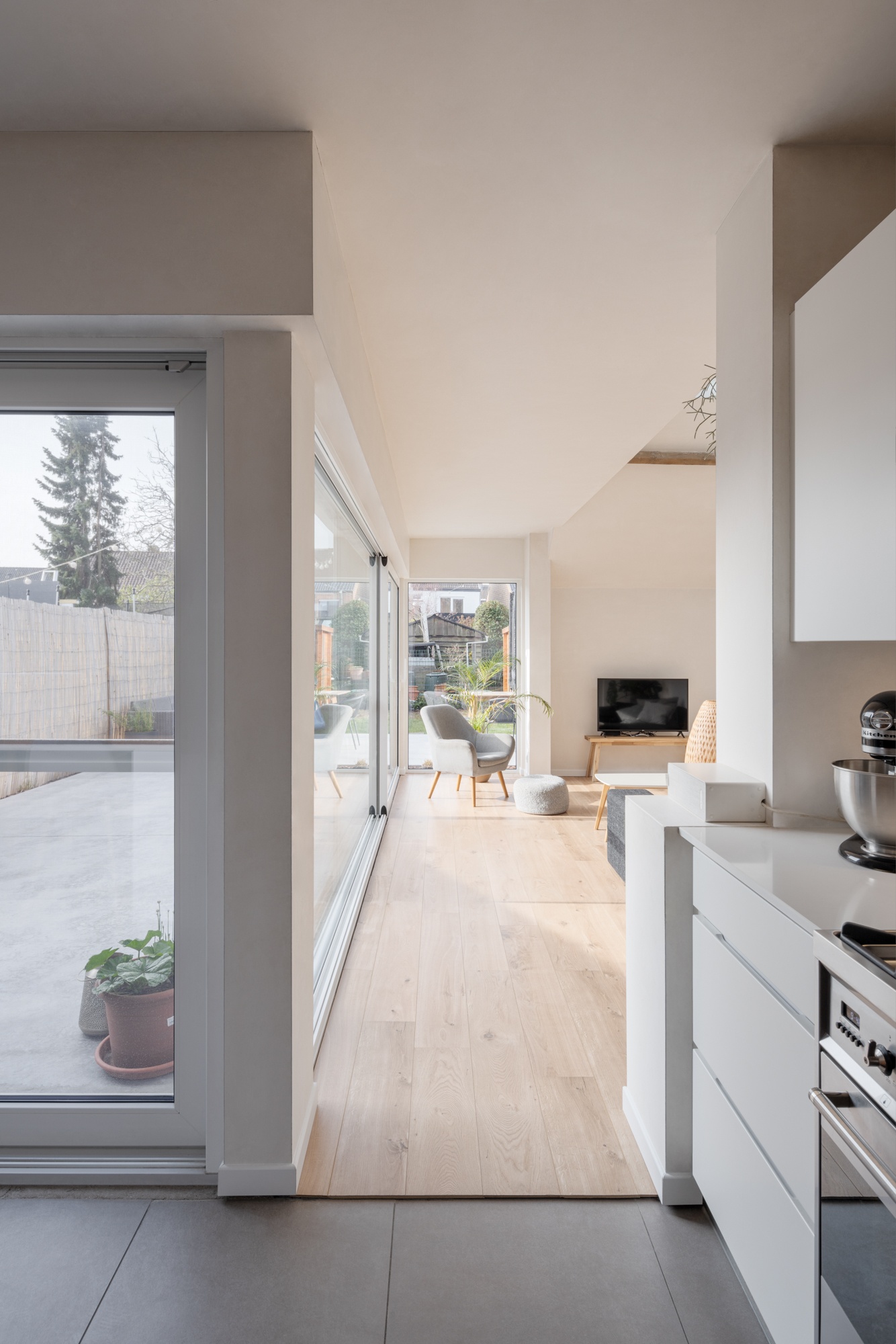 Groot vast raam met witte aluminiumprofielen in een lichte, gerenoveerde gesloten bebouwing, met zicht op de leefruimte, keuken en terras.