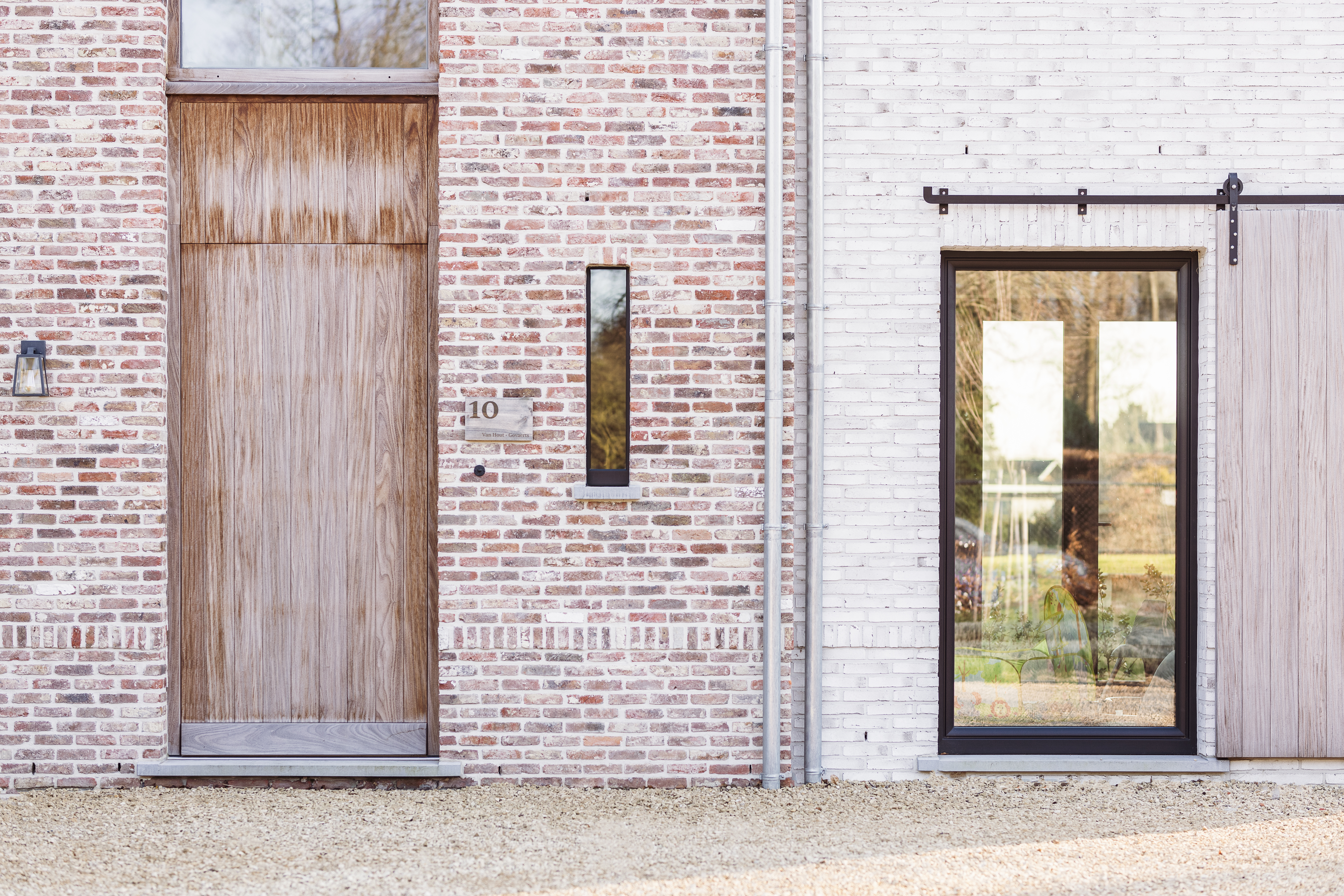 Bakstenen gevel met een hoge houten deur, een smal verticaal raam en een matzwart, rond schuifraam met houten luik.