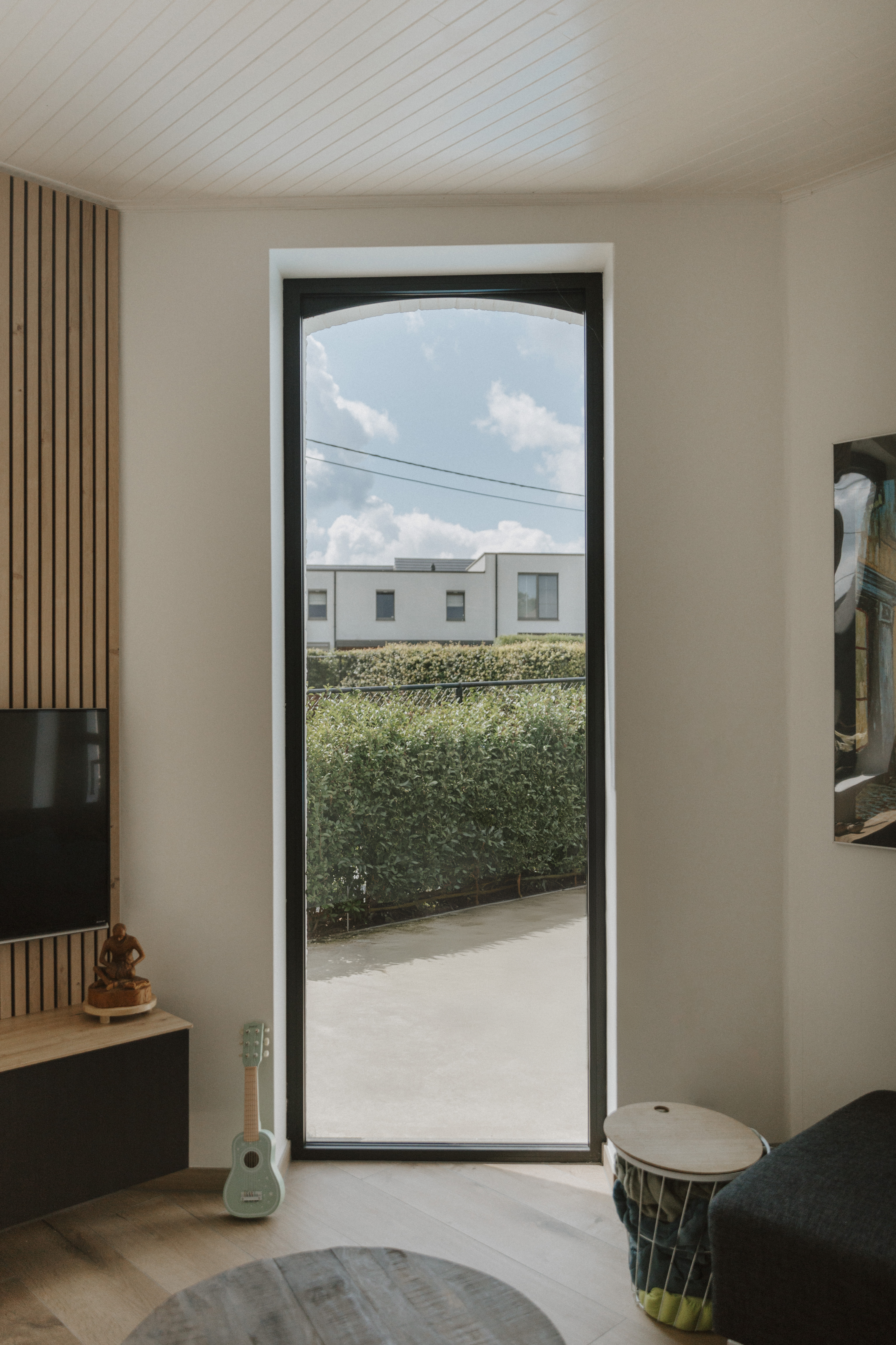 Vast raam met slank, diepzwart raamprofiel in een interieur, met zicht op een haag, een verharde buitenruimte en een woning.
