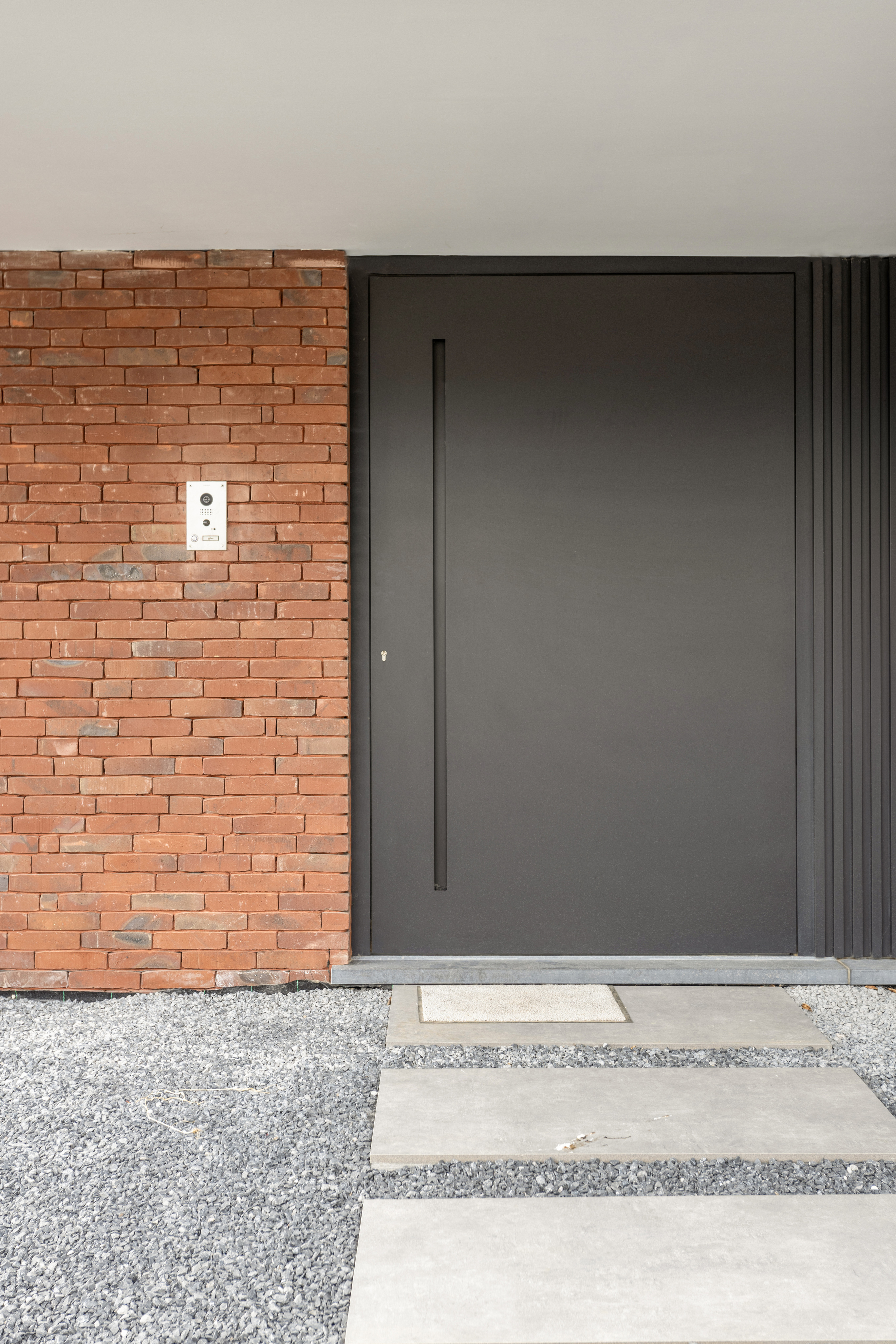 Zwarte aluminium paneeldeur van ALU Koncept in een karaktervolle, moderne stijl, geplaatst in een rode bakstenen gevel met een pad in betontegels.