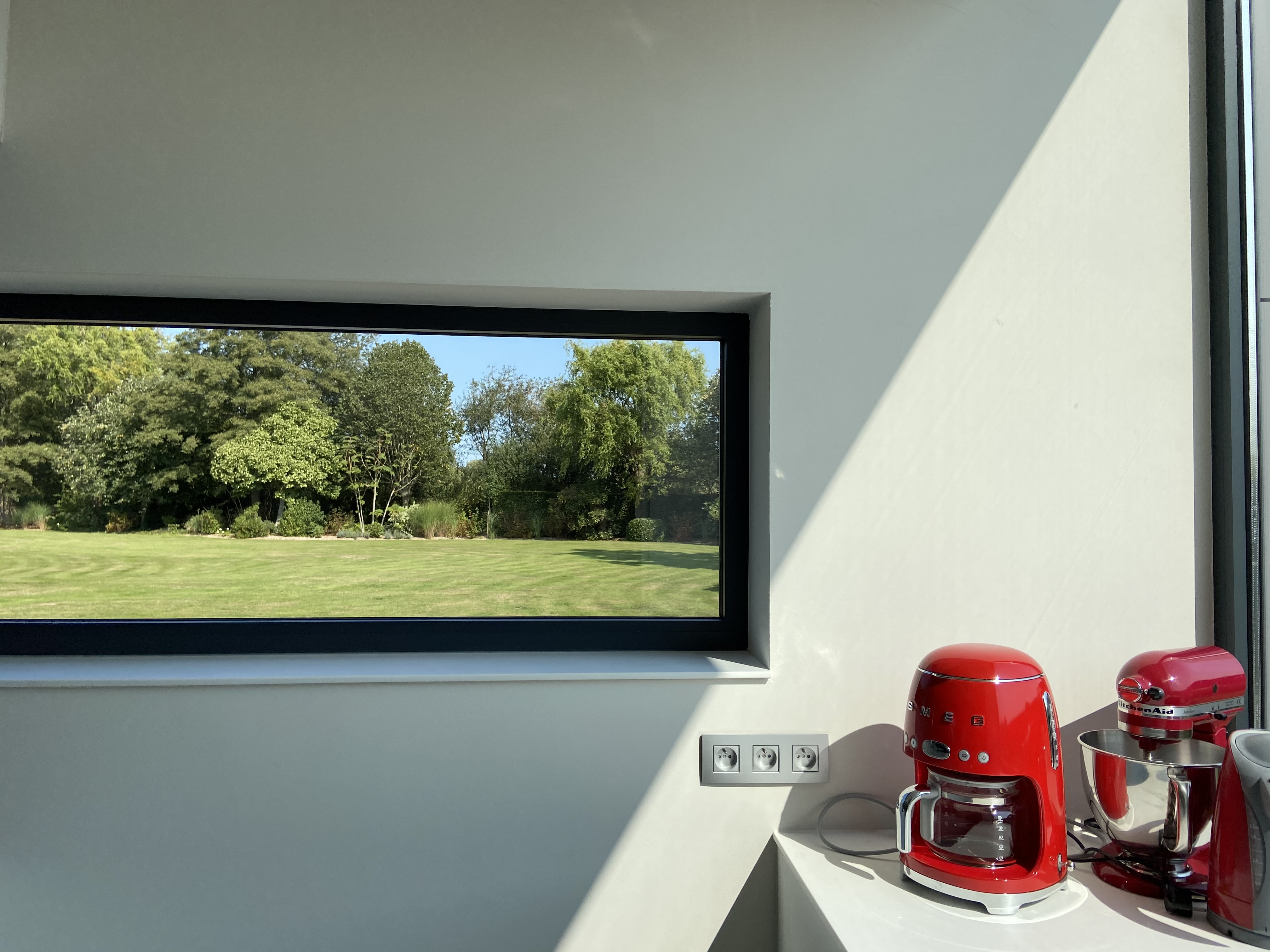 Een zwart raam in een moderne keukenwand kijkt uit op een ruime tuin; rode keukenapparaten staan op het werkblad.