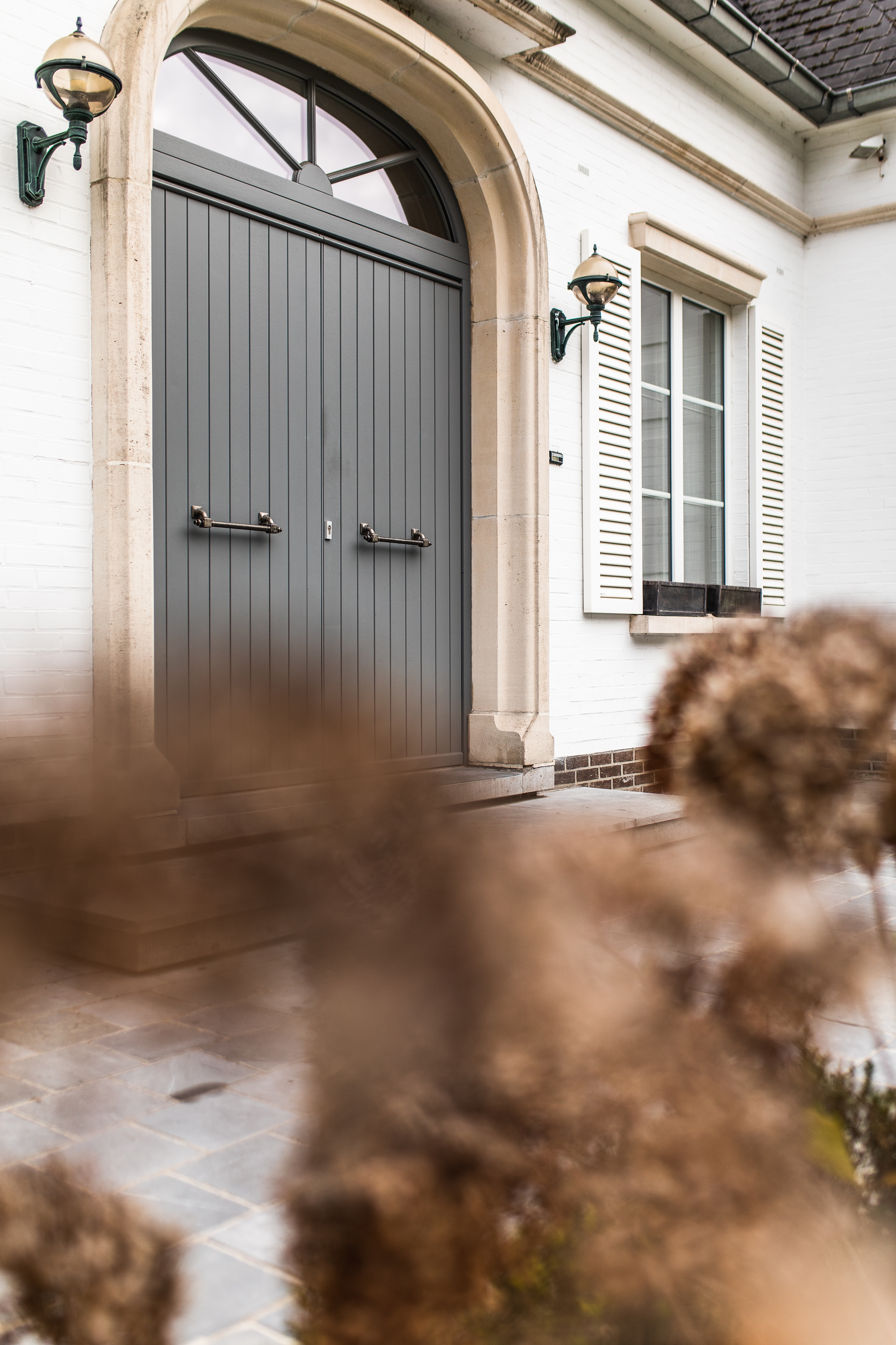 Witte gevel van een gerenoveerde vrijstaande woning met verfijnde klassieke details, een grijze gebogen deur, witte luiken en een terras in natuursteen.