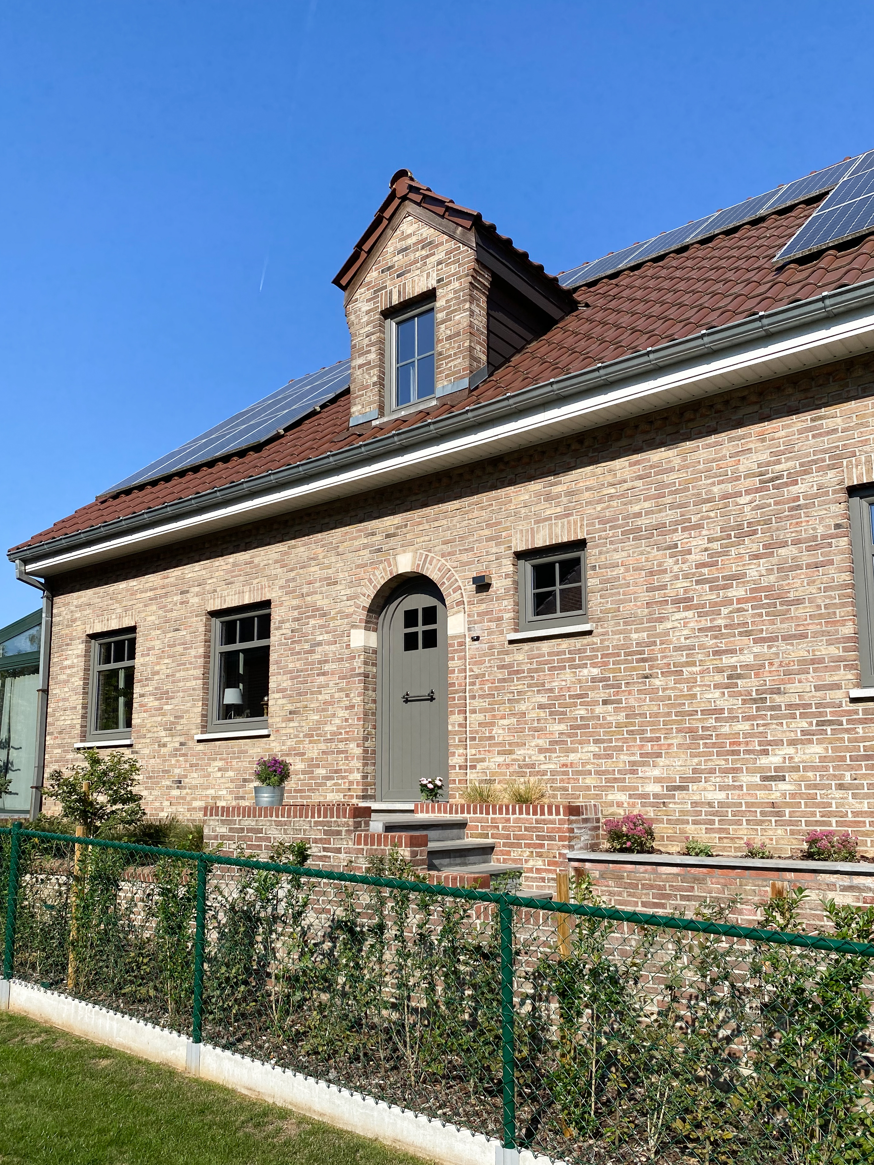 Gerenoveerde vrijstaande bakstenen woning met een semi-beglaasde Kwartsgrijze NERF-deur, meerdere dakramen en een kleine omheinde voortuin.