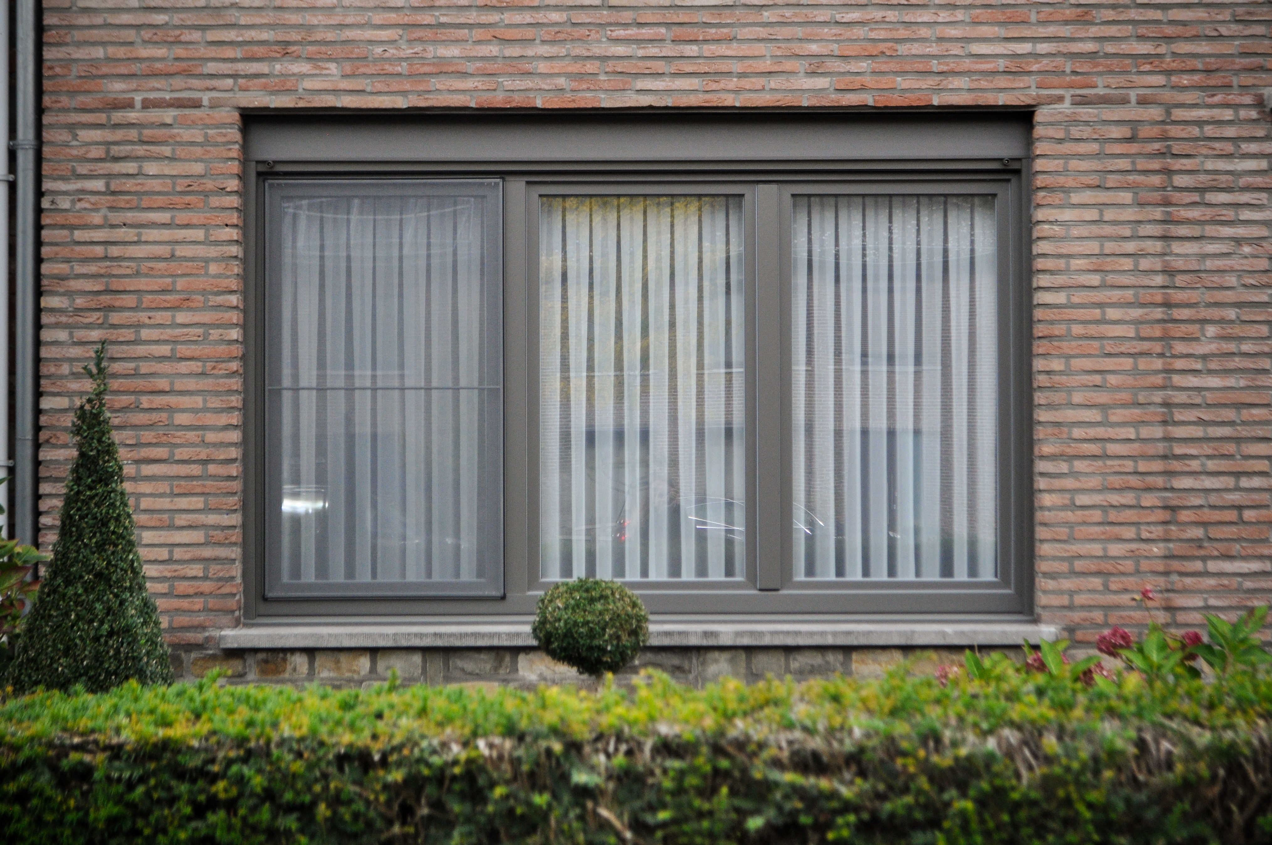 Kwartsgrijs NERF-raam met drie verticale delen in een gemetselde gevel, met gesloten raamdecoratie en een lage haag op de voorgrond.