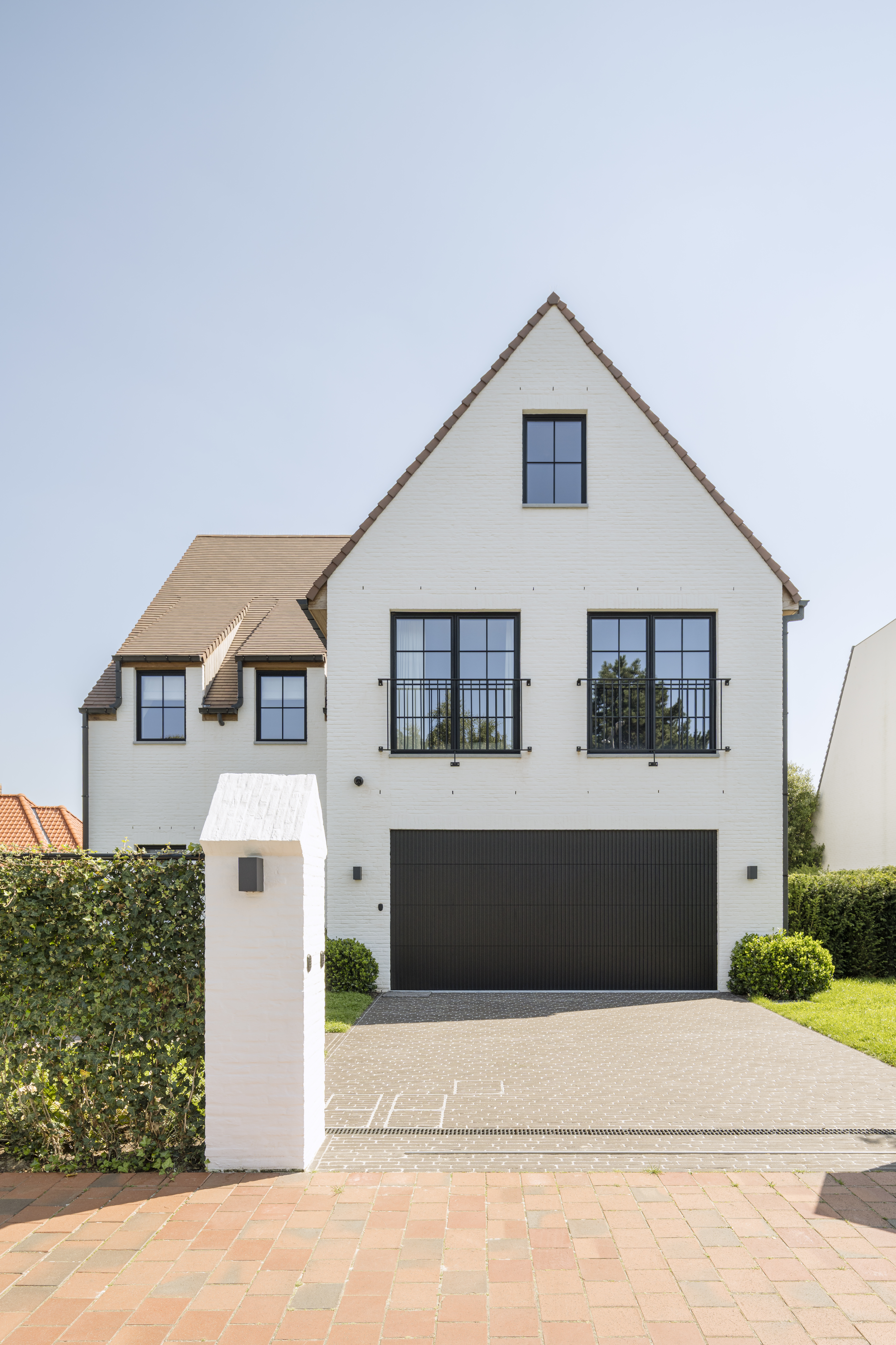 Moderne witte woning met een zadeldak, grote ramen, donkere garagepoort, geplaveide oprit en groene hagen.