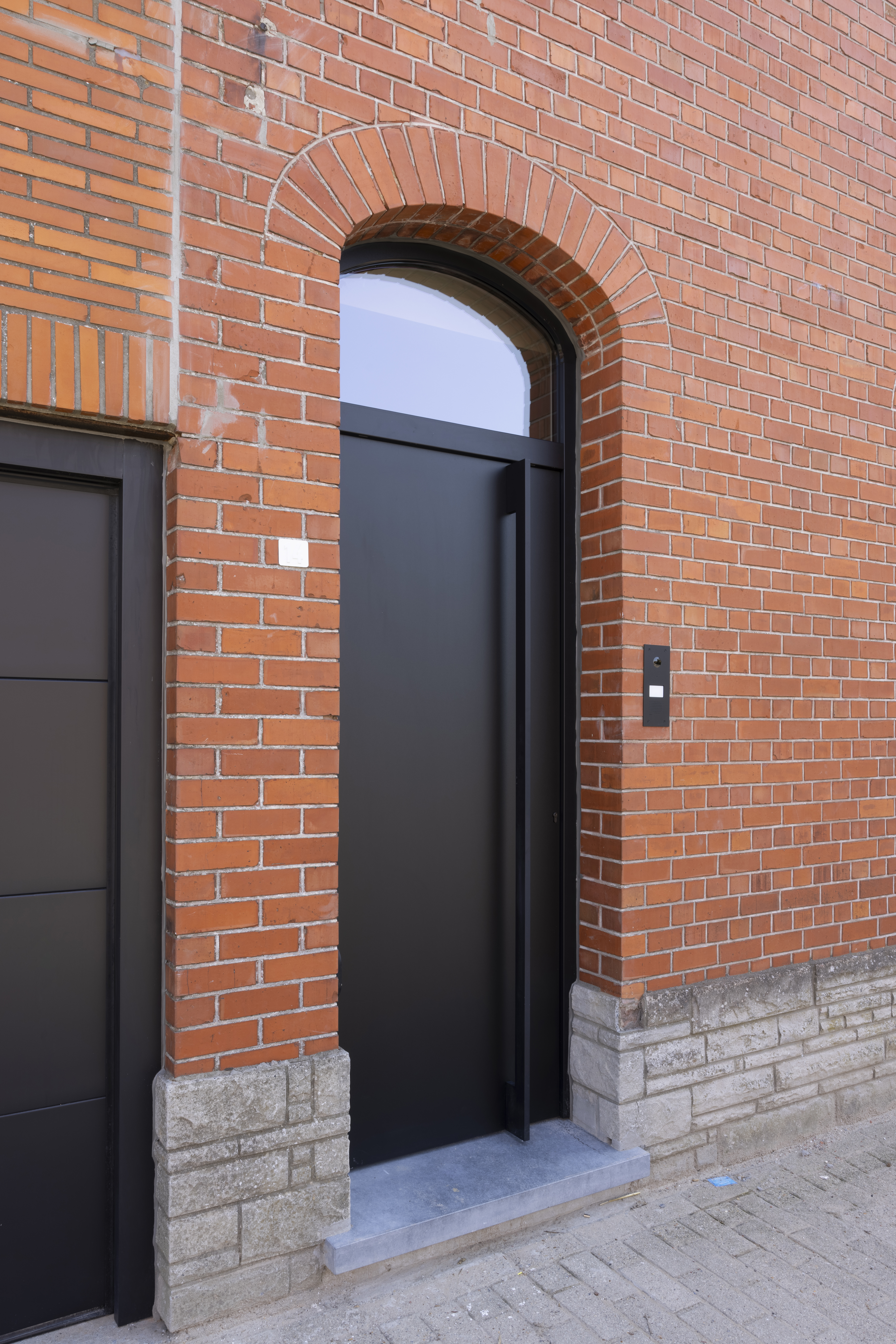 Mat zwarte PVC Komfort PLUS-deur met bovenlicht en zijpaneel in een rode bakstenen gevel aan de inkom.