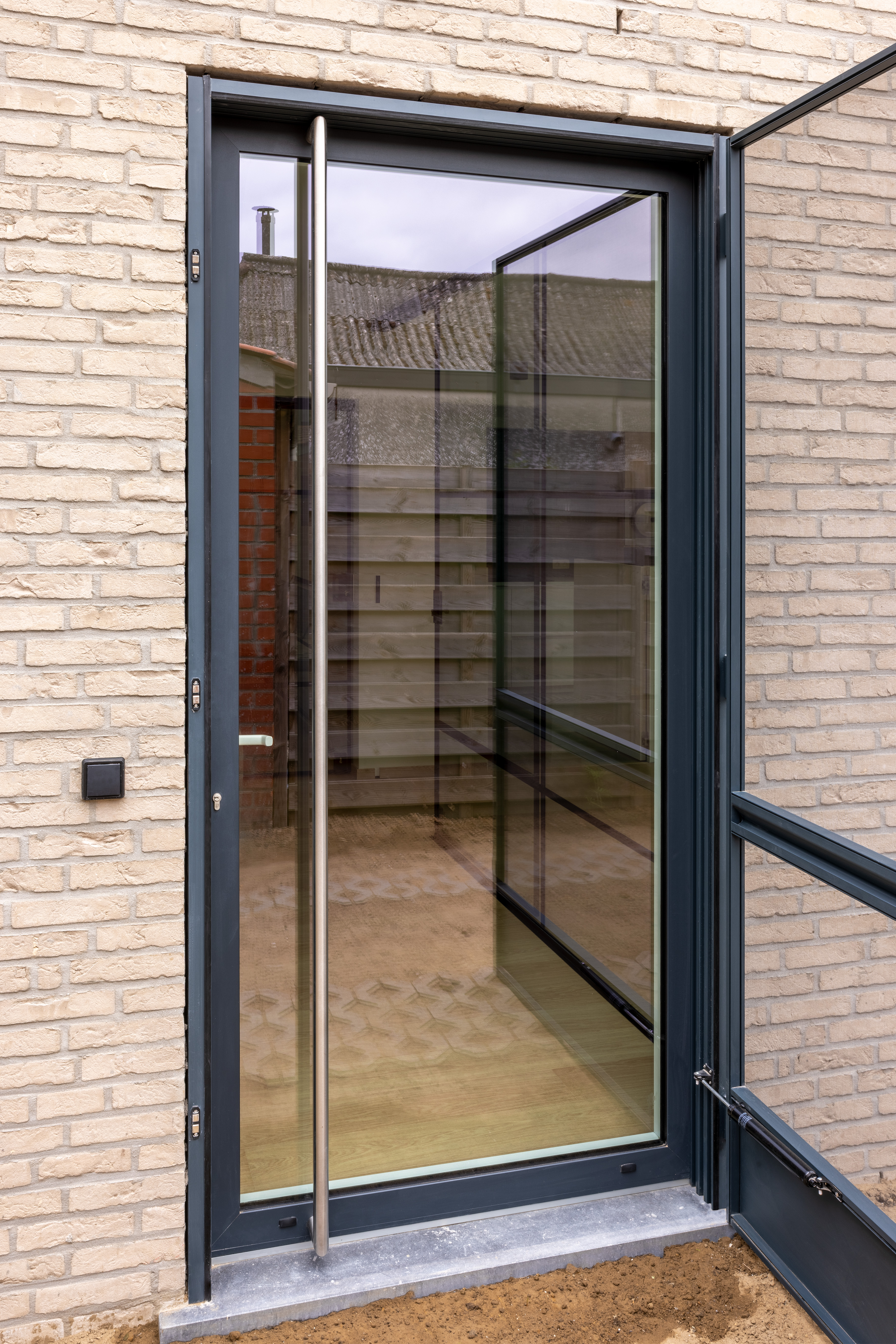 Moderne beglaasde aluminiumdeur in antracietgrijs met vliegendeur, geopend in de bakstenen gevel van een gerenoveerde gesloten bebouwing.