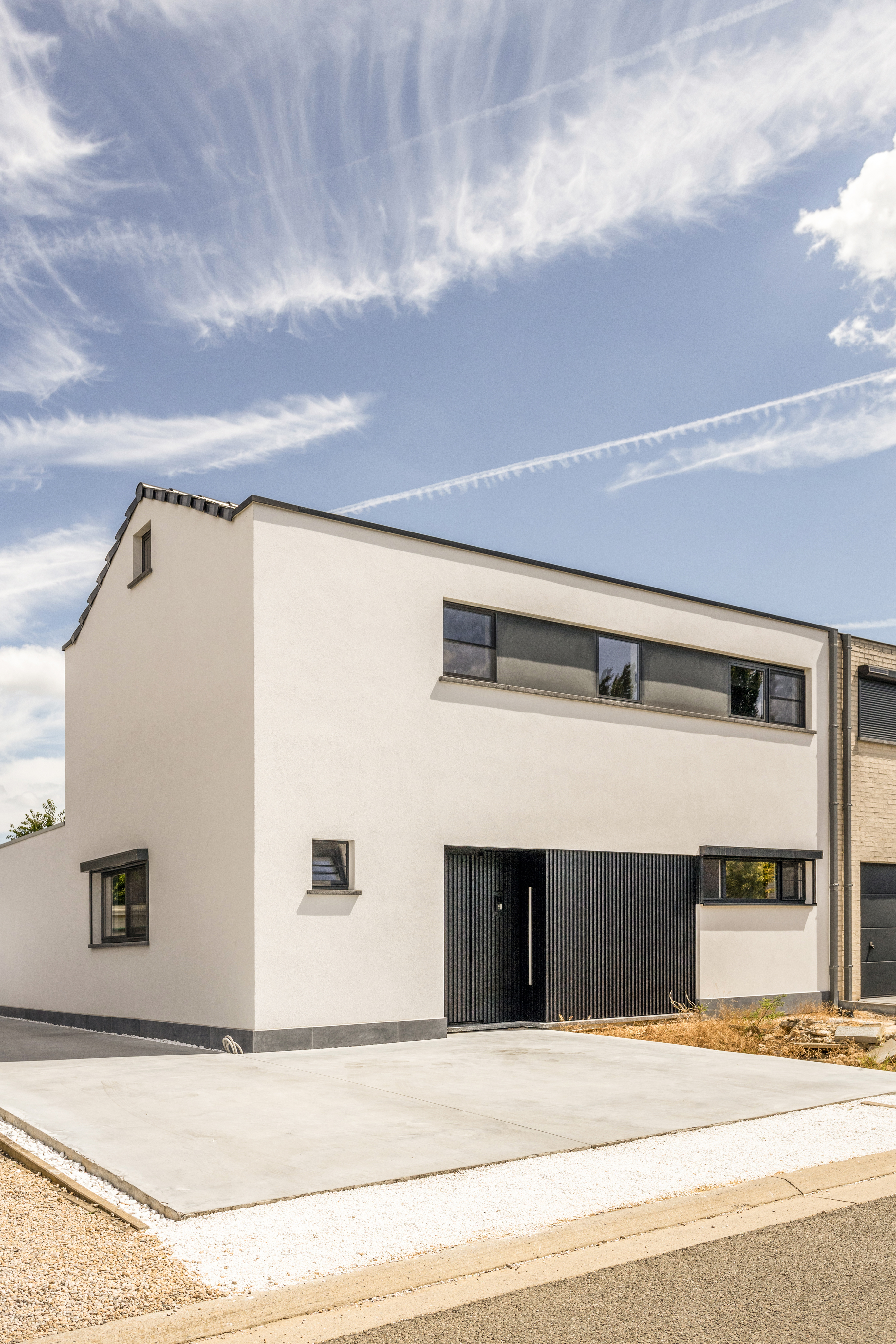 Witte woninggevel met lange ramen, een donkere inkomdeur en een zwarte garagepoort aan een brede oprit onder een blauwe hemel.