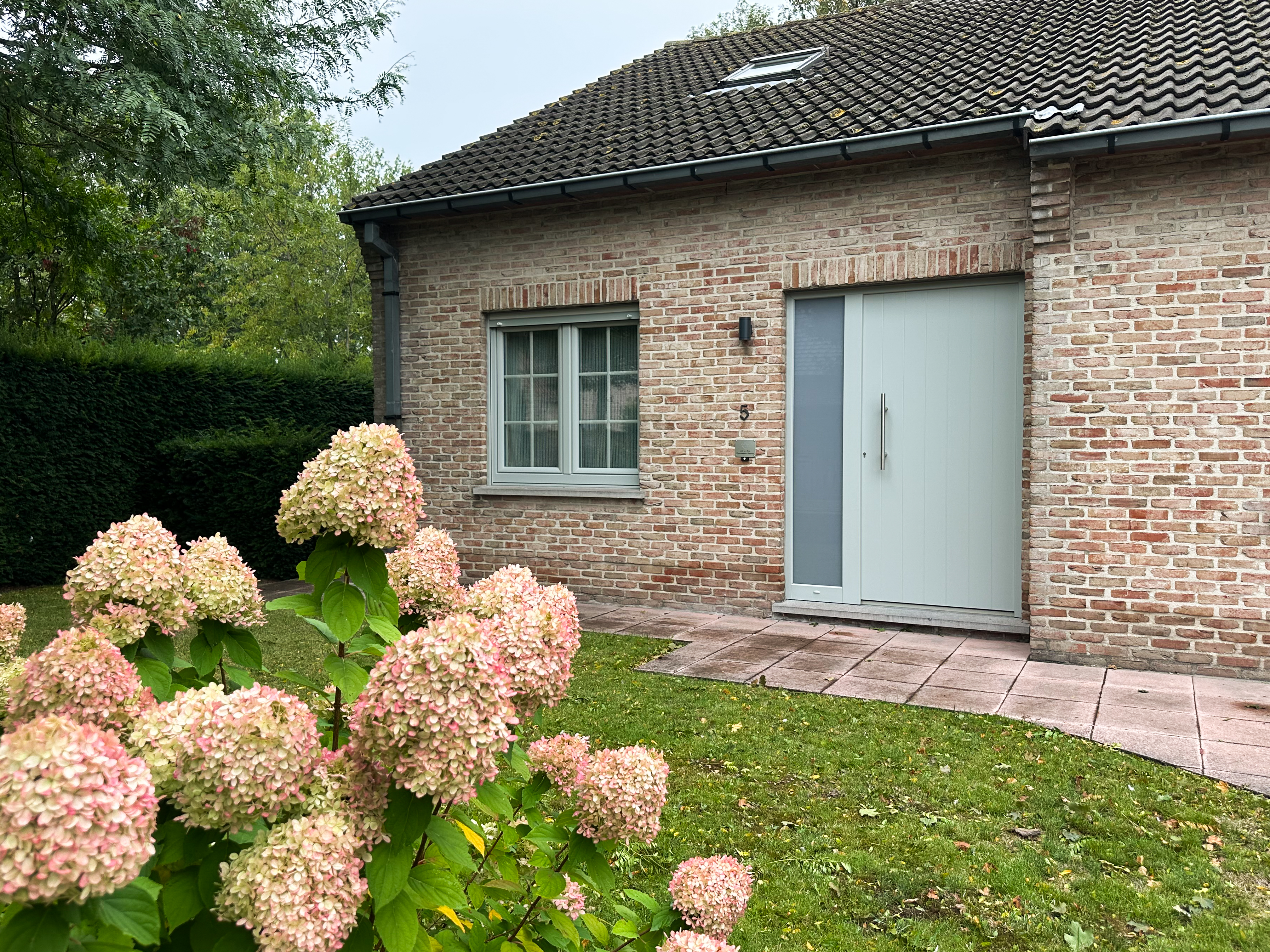 Bakstenen gevel met een agaatgrijze PVC-deur met smal zijlicht en een dubbel draai-kipraam, omgeven door gazon en hortensia’s.