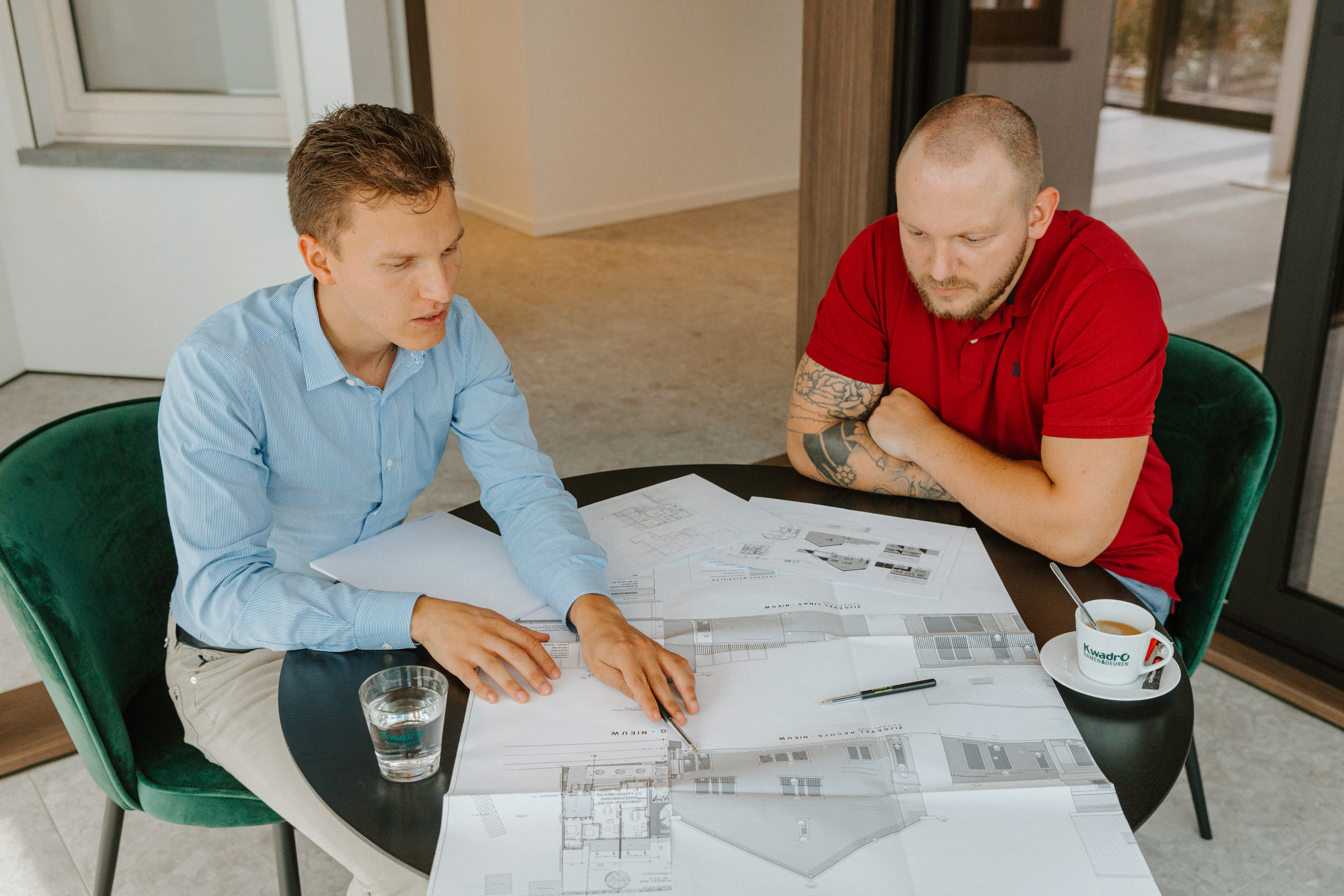 Deux hommes examinent ensemble des plans de construction autour d’une table dans un intérieur lumineux, avec un verre d’eau et une tasse à côté.