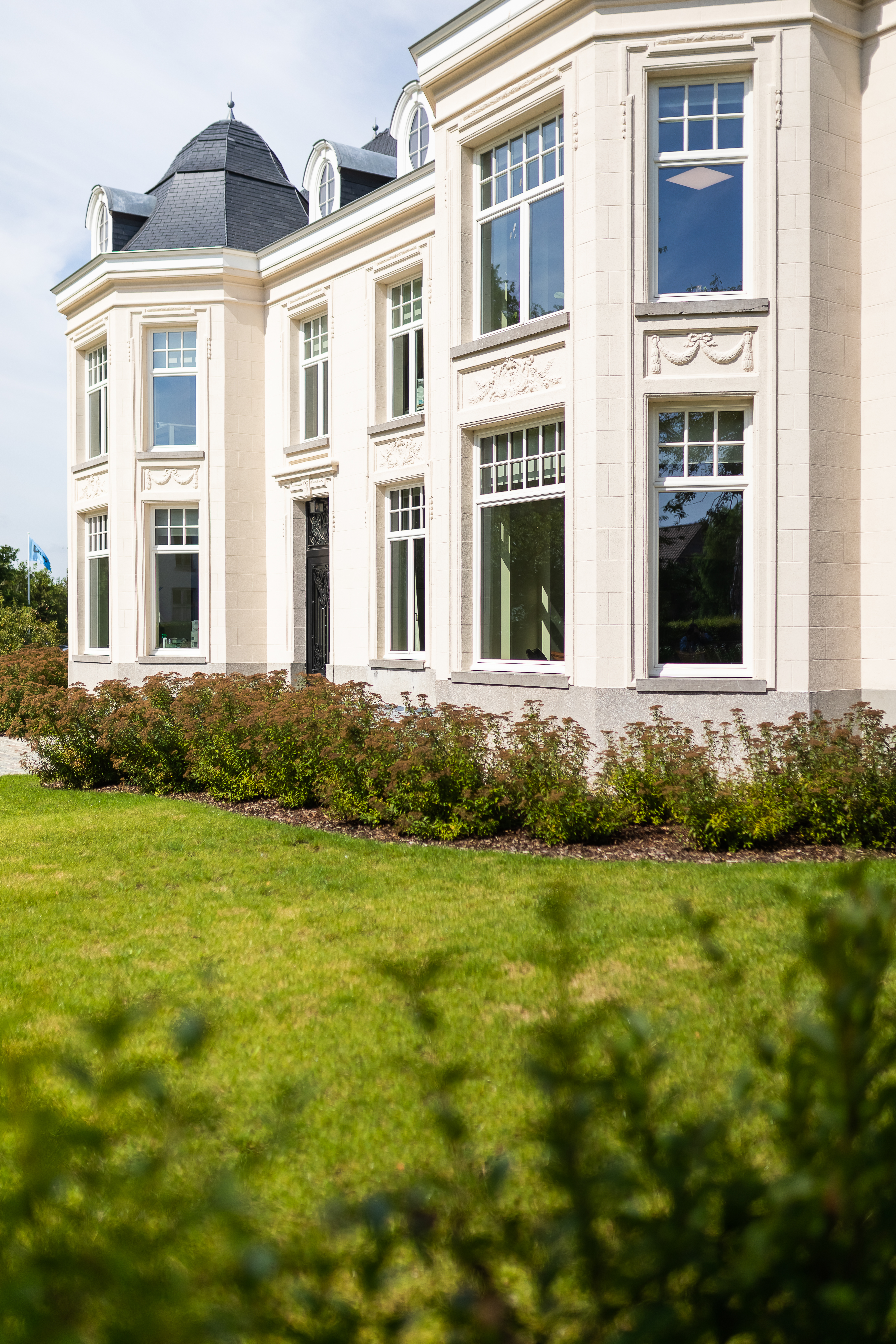 Witte gevel in verfijnde klassieke stijl van een gerenoveerde vrijstaande woning, met hoge ramen, sierlijke raamprofielen, een torentje en een verzorgde voortuin.