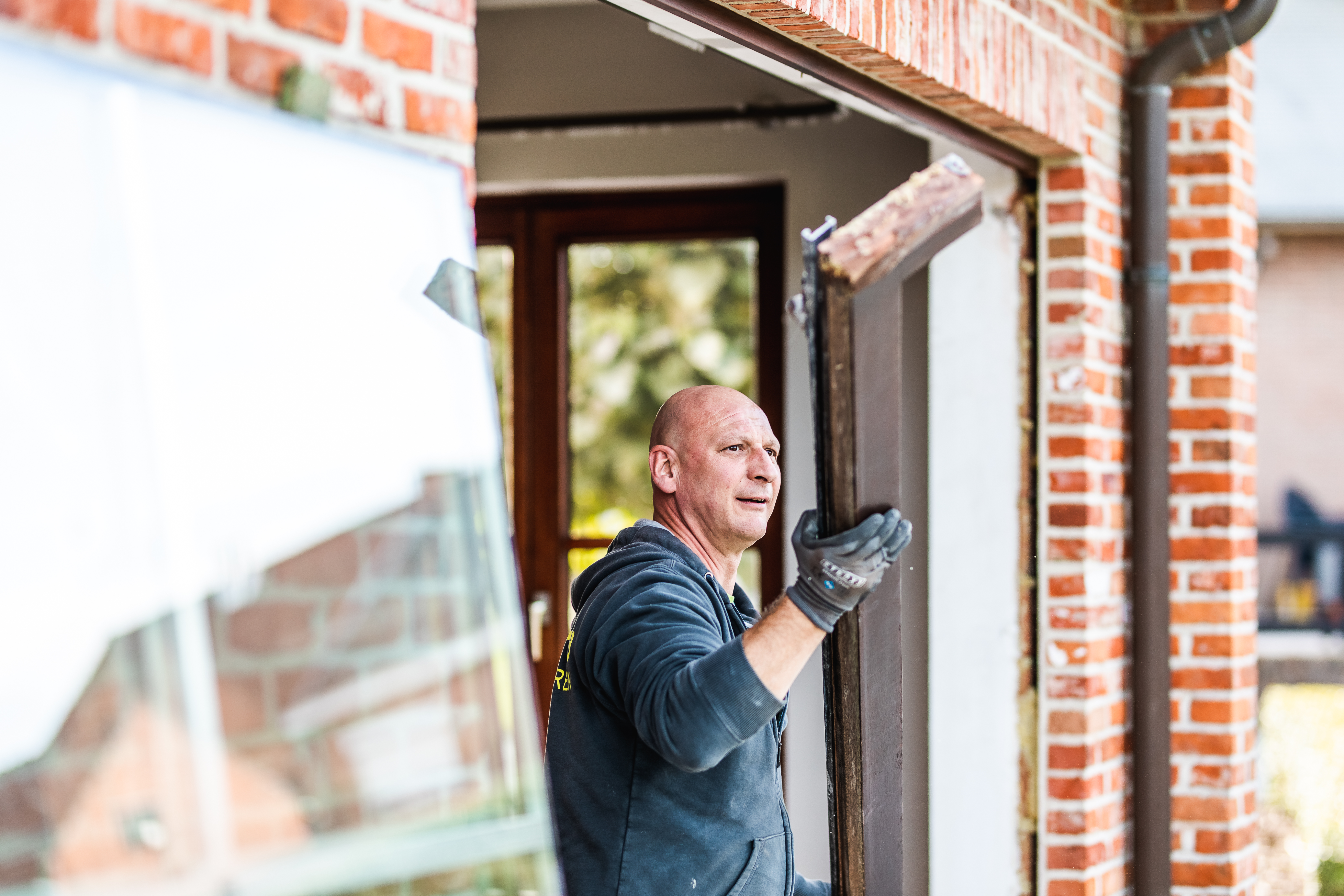 Een persoon in werkkledij en handschoenen werkt aan een deurkader in een bakstenen gevel en houdt daarbij een donker onderdeel vast.
