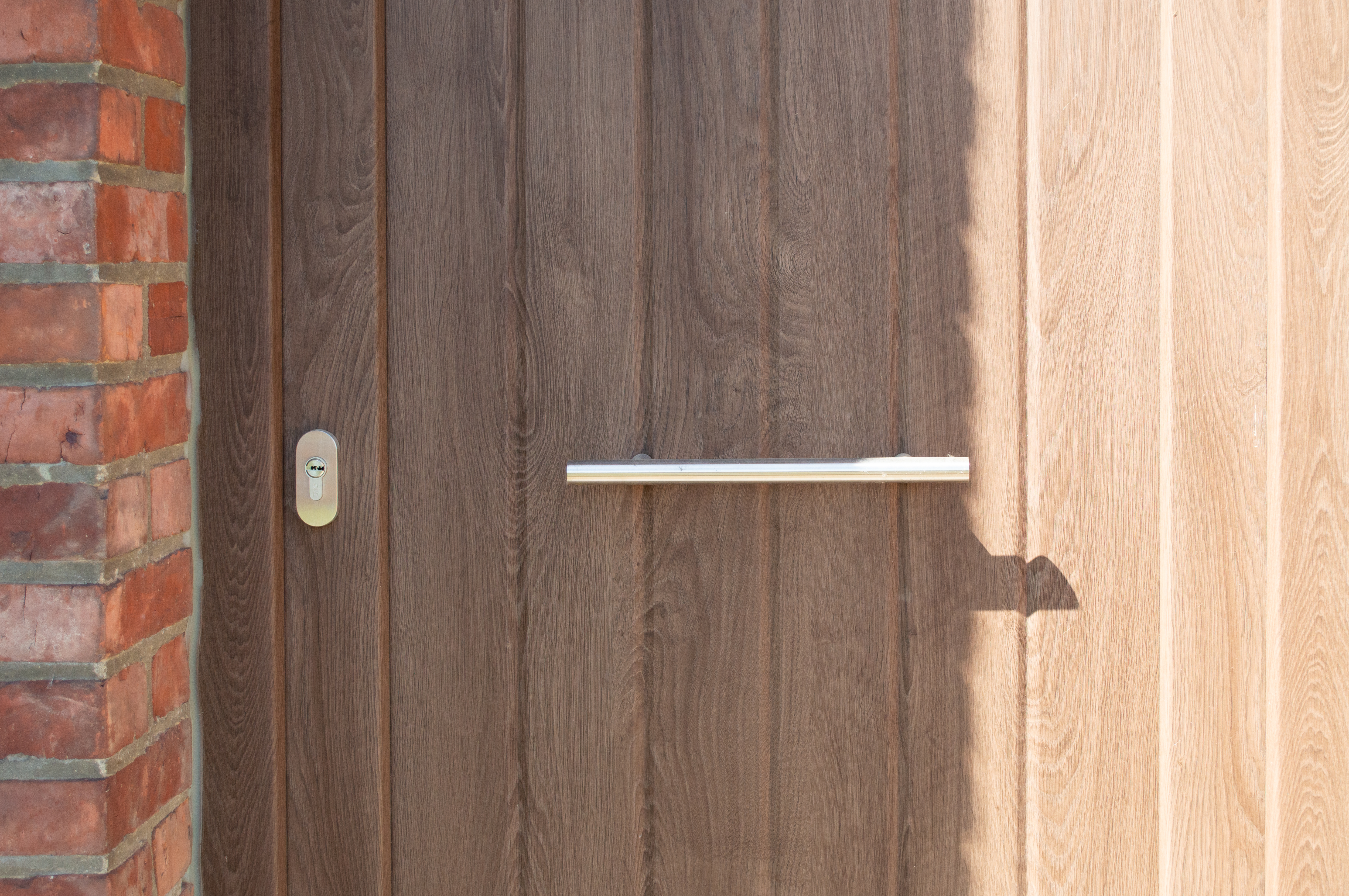 Donkerbruine pvc-deur met een houtstructuur in Woodec Donkere eik, een horizontale metalen handgreep en een bakstenen muur ernaast.