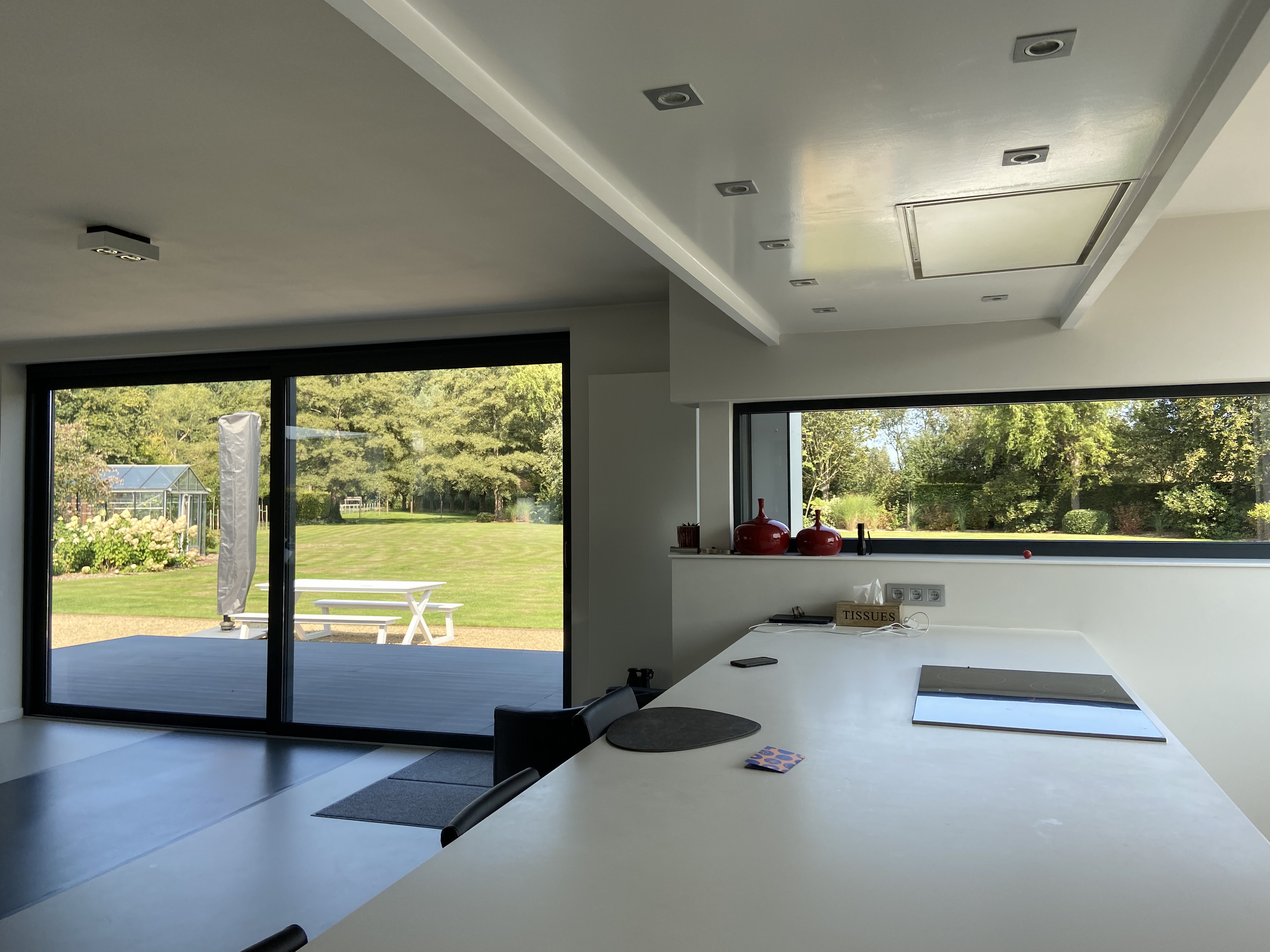 Moderne keuken in een vrijstaande woning met een groot zwart schuifraam naar de tuin, een kookeiland en een brede horizontale raampartij.