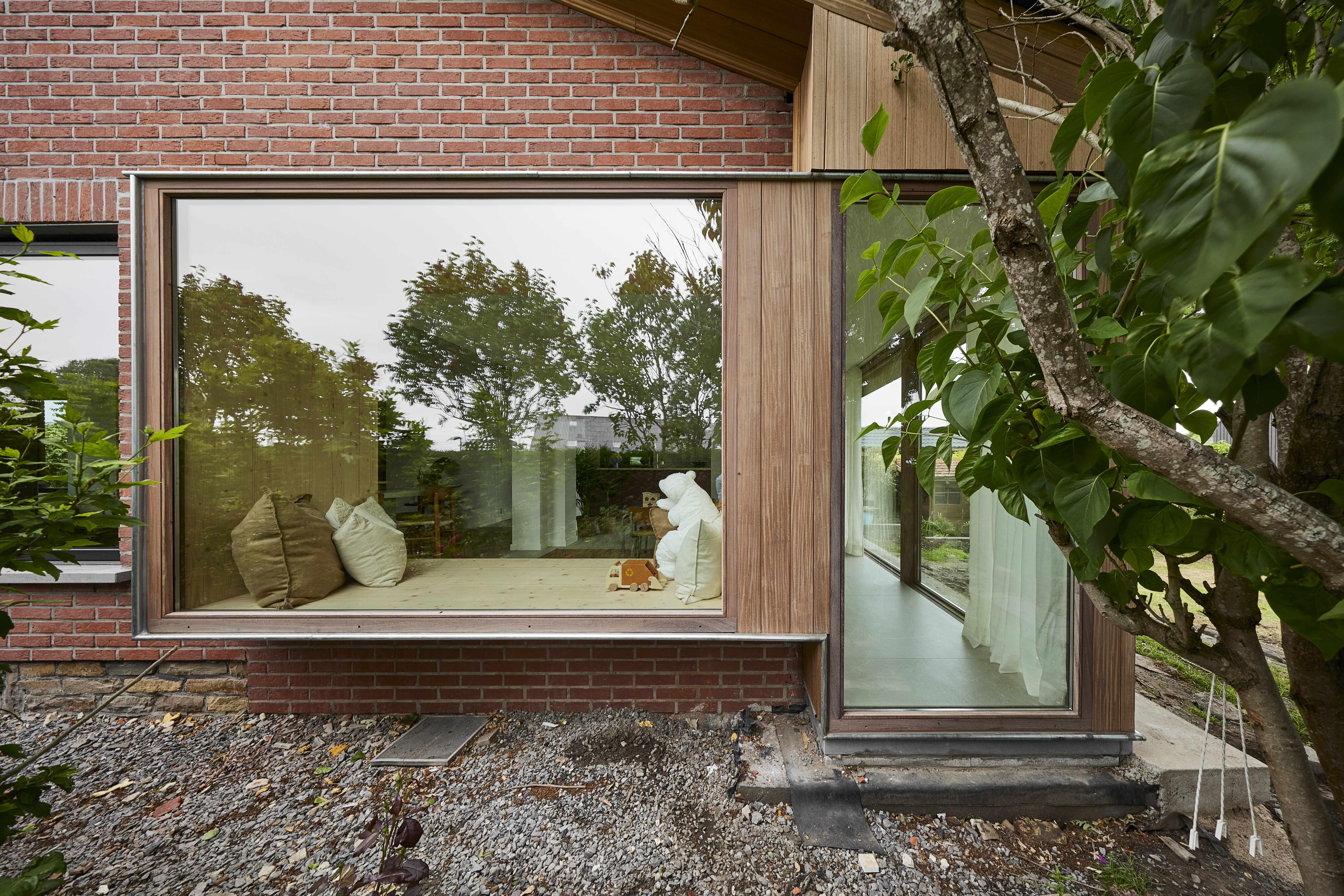 Modern vast raam met een natuurlijke houten afwerking in een rode bakstenen gevel, naast een smal glazen zijpaneel en een groene buitenruimte.