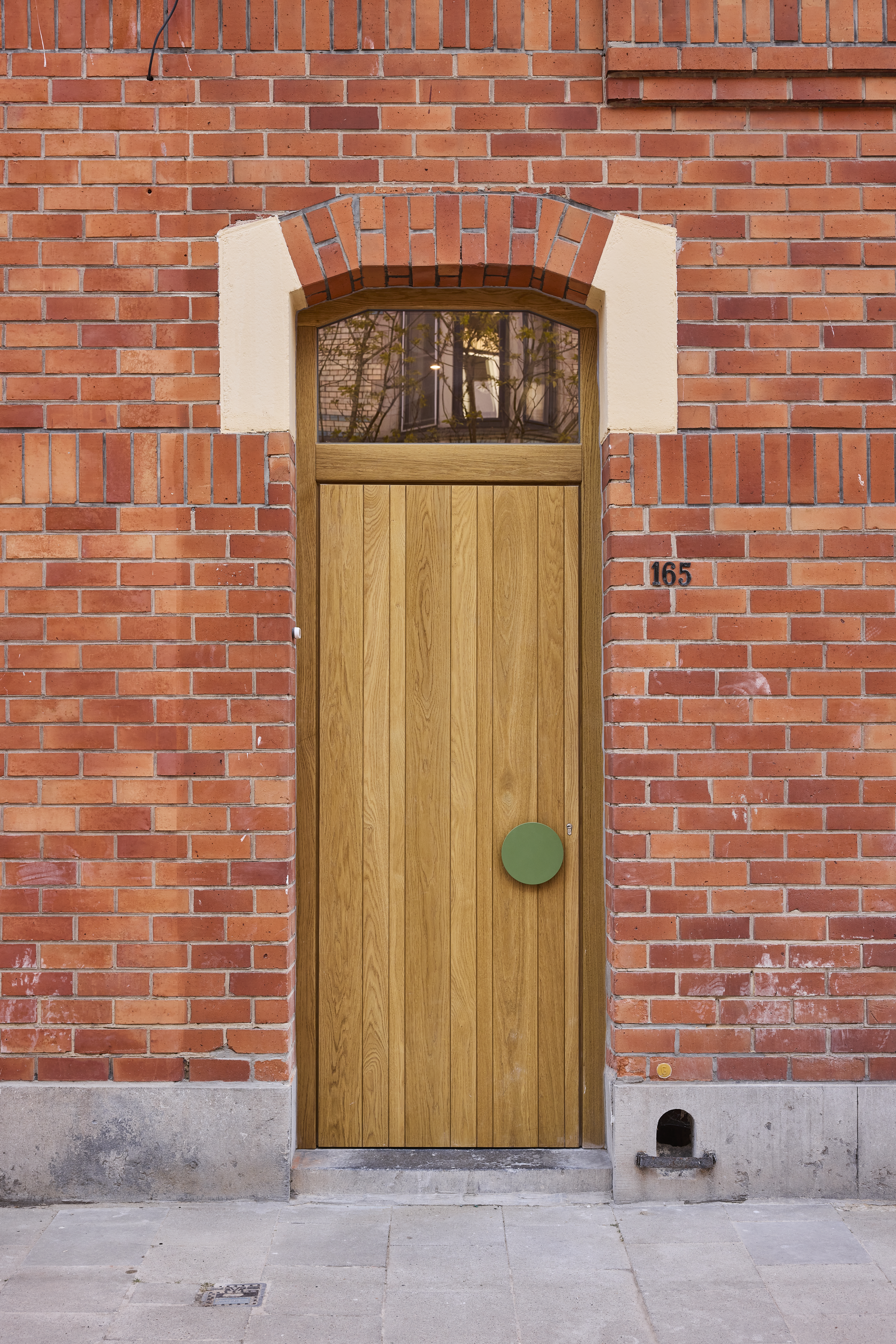 Eikenhouten deur met bovenlicht in een gevel van rode baksteen, met een groene deurknop en huisnummer 165.