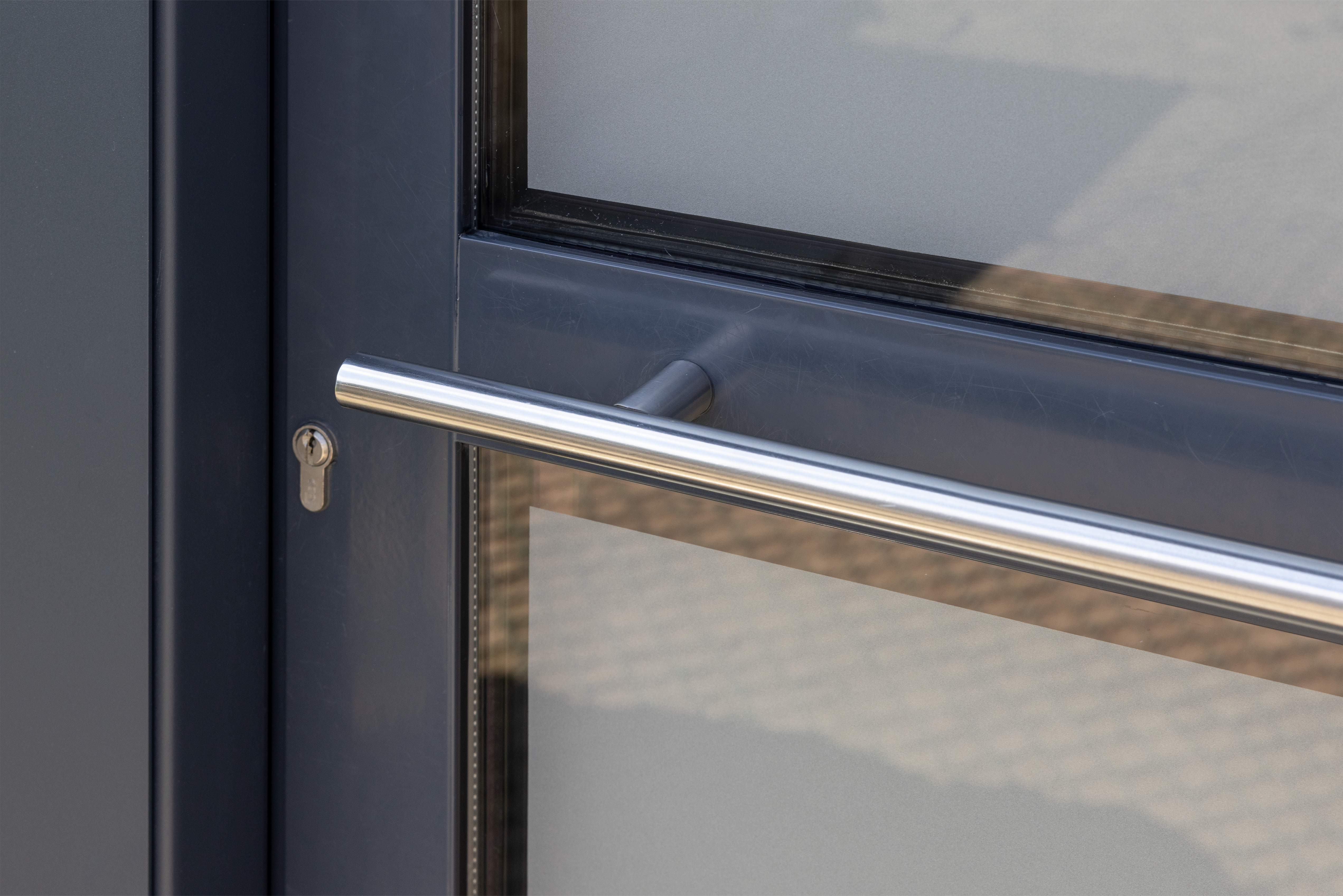 Close-up van een antracietgrijze deur met glasvlakken, een lange horizontale metalen handgreep en cilinderslot, als tijdloos detail bij een renovatie.