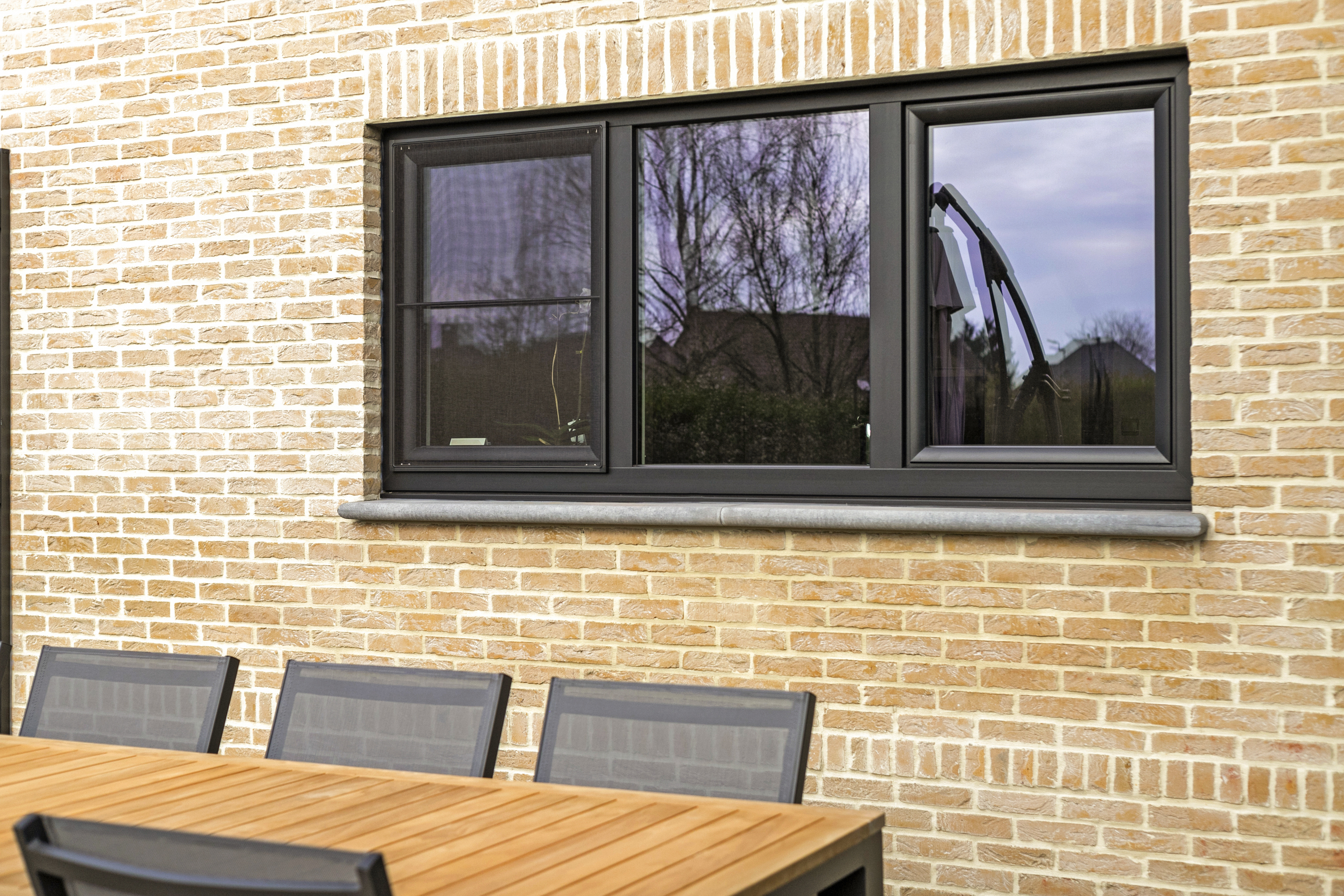 Matzwart dubbel raam met draai-kipraam en vliegenraam in een warme, landelijke bakstenen gevel, boven een houten tuintafel.