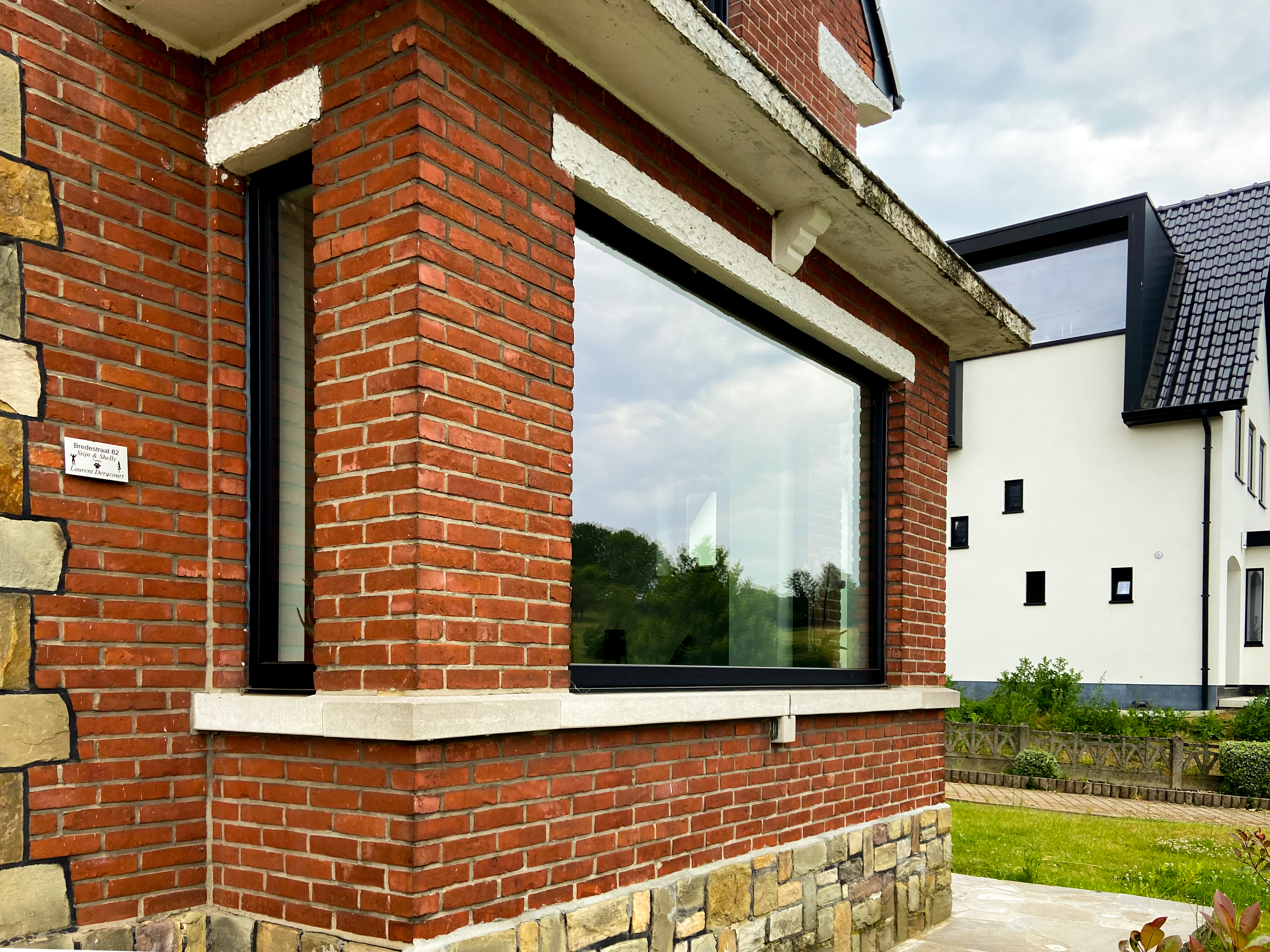 Groot matzwart raam in de rode bakstenen gevel van een gerenoveerde vrijstaande woning, met een aangrenzende witte gevel en tuin.