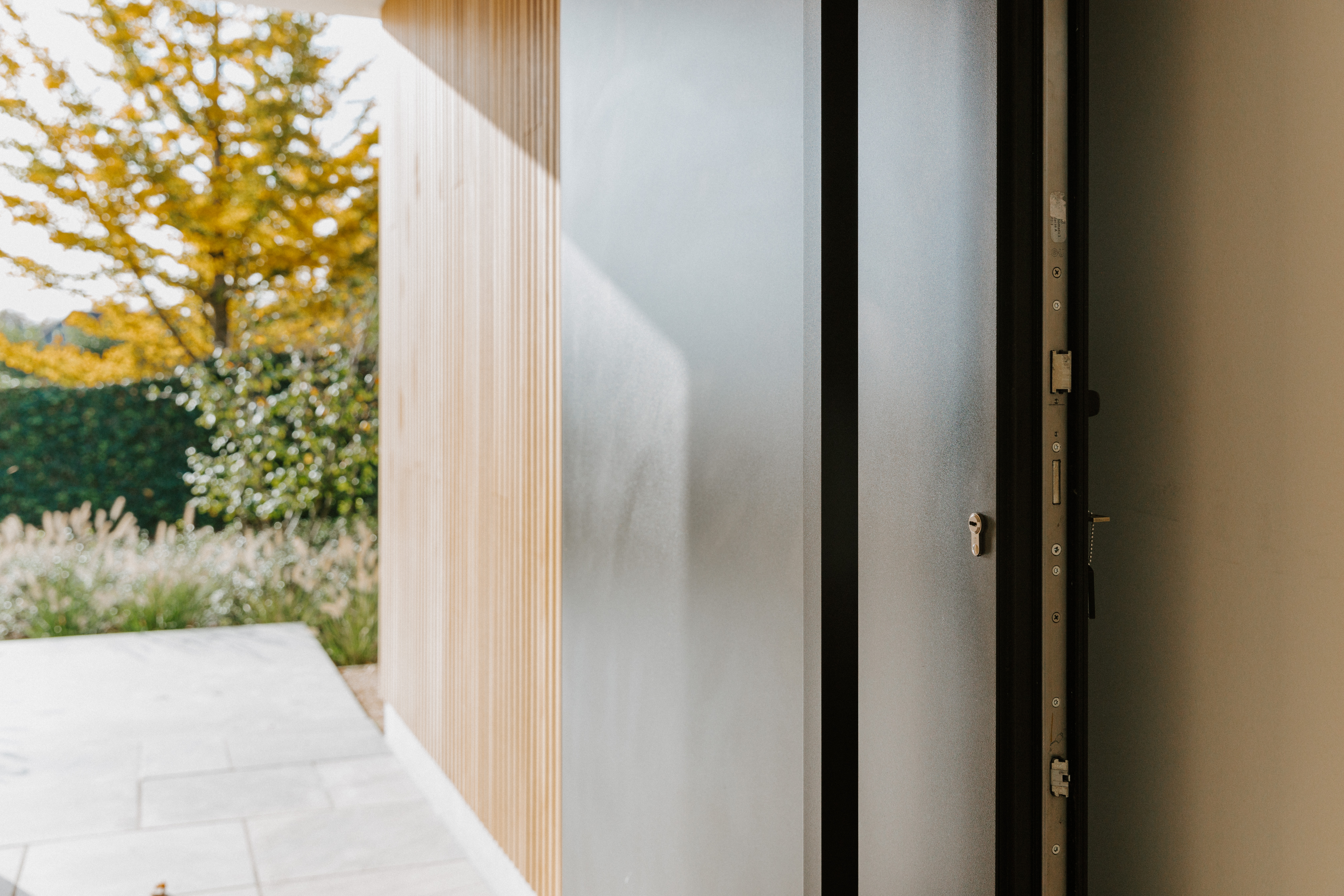 Moderne zwarte aluminium voordeur met paneel, openstaand naast een lichte gevel met houten verticale bekleding en een betegeld terras.