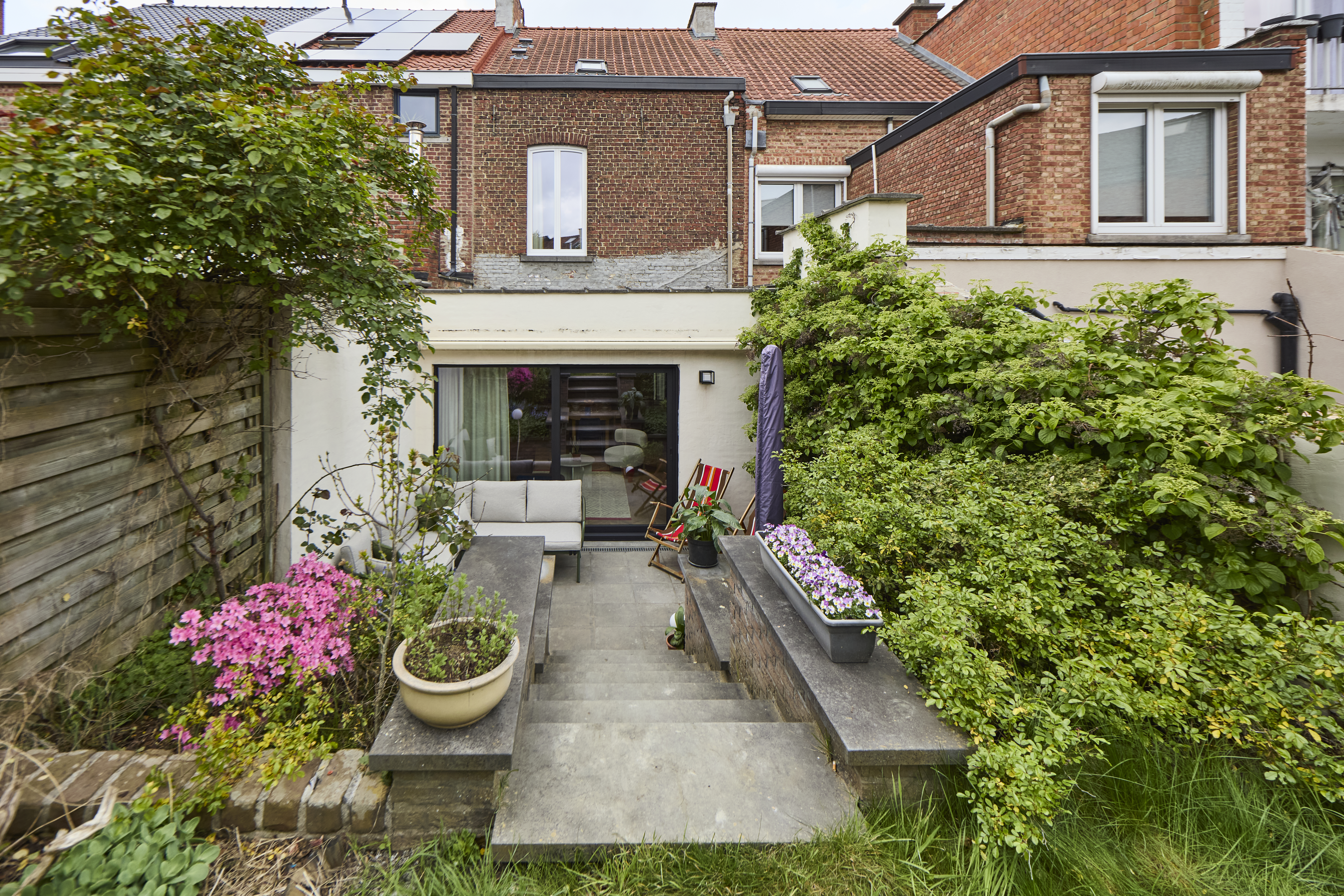Een zwart ALU Visoglide Monorail-schuifraam in Signaalzwart 9004TC aan de achtergevel geeft uit op een betegelde tuin met bloembakken, struiken en een houten afsluiting.