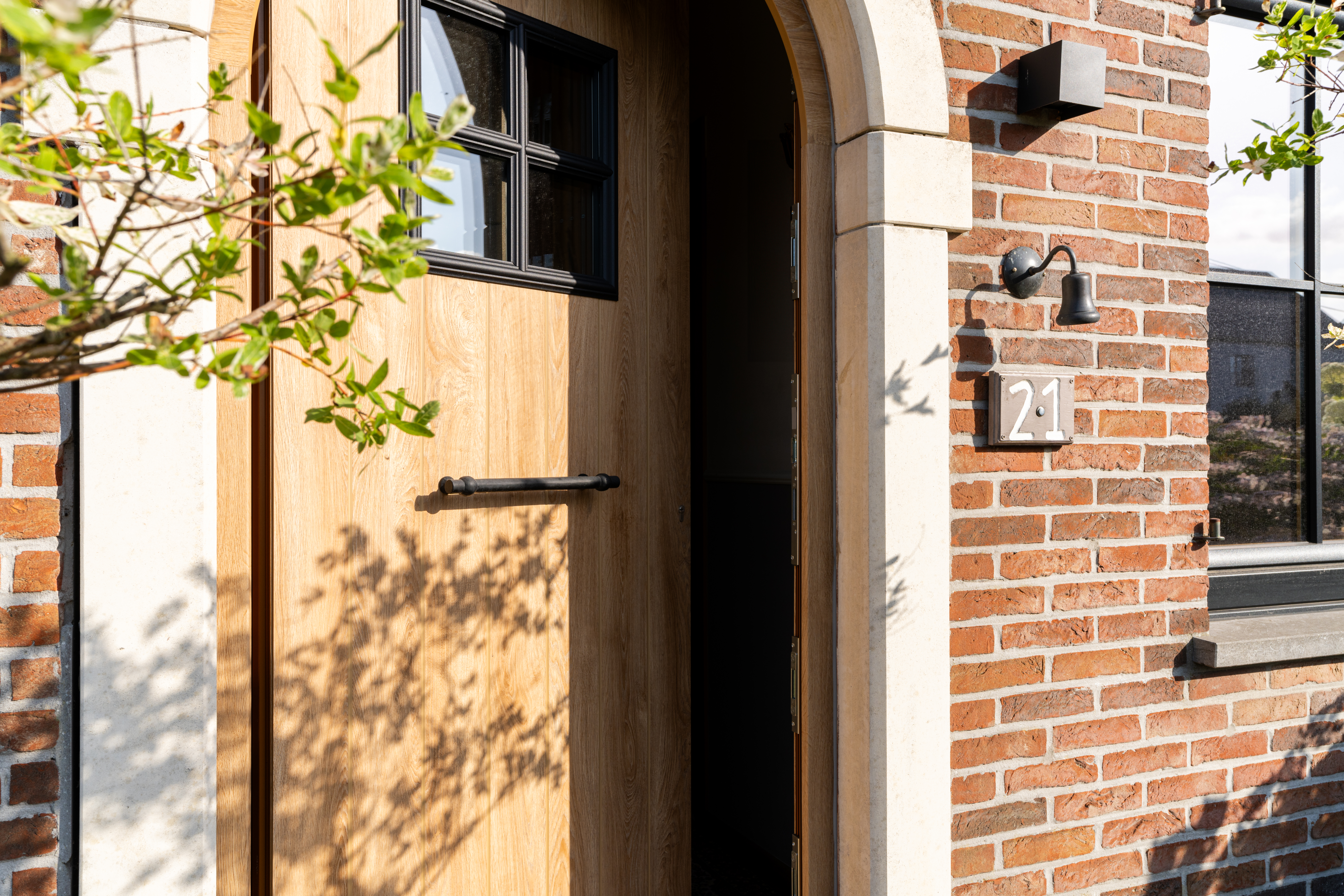 PVC-voordeurpaneel met Woodec Eik Natuur-afwerking, kleine glasvlakken, zwarte handgreep en brievenbus in een bakstenen gevel naast een zwart raam.